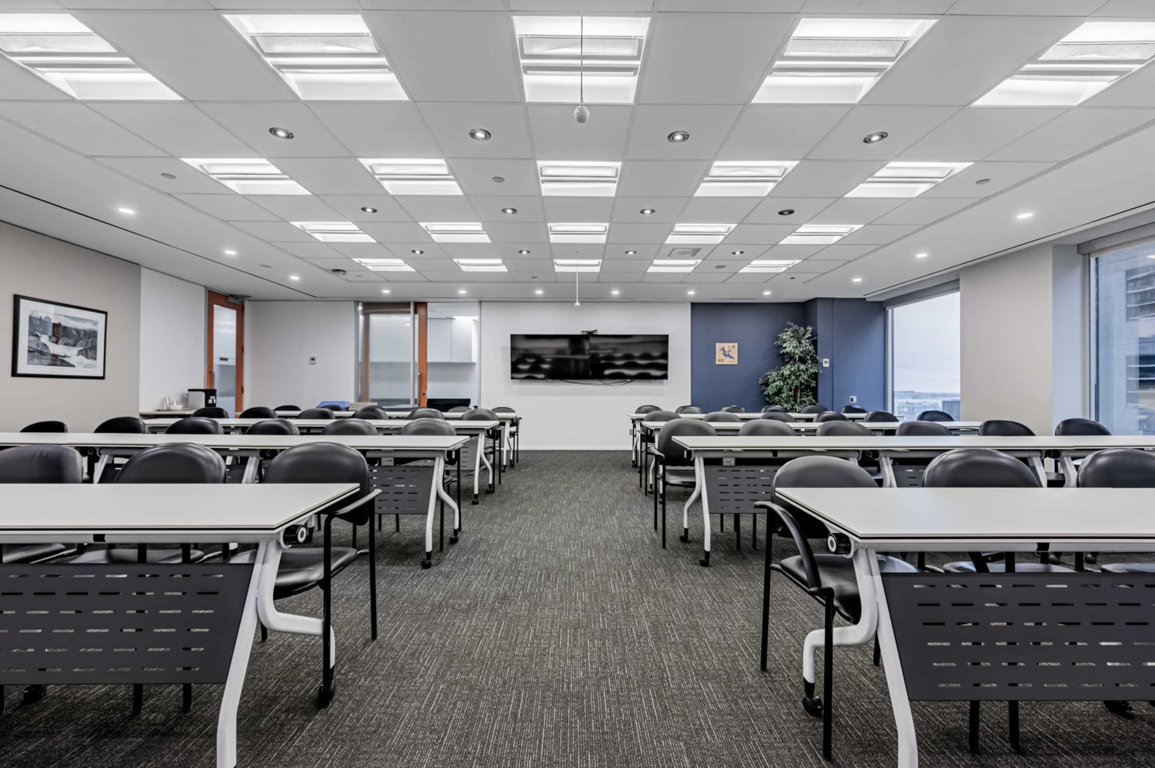 A modern conference room features rows of tables and chairs, large windows, and a wall-mounted screen.