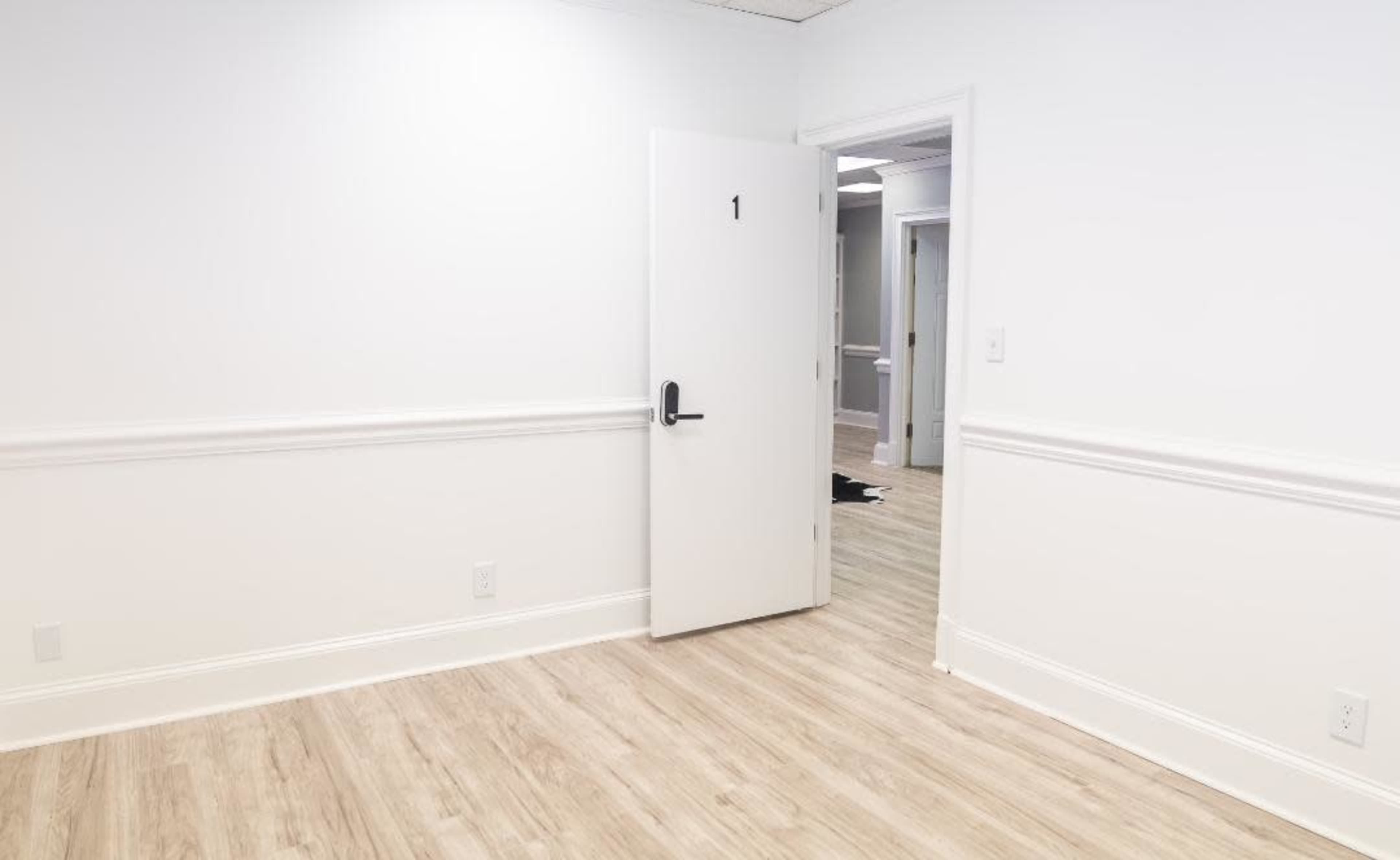 A white room with light wooden flooring features an open door numbered "1" and a partially visible hallway beyond.