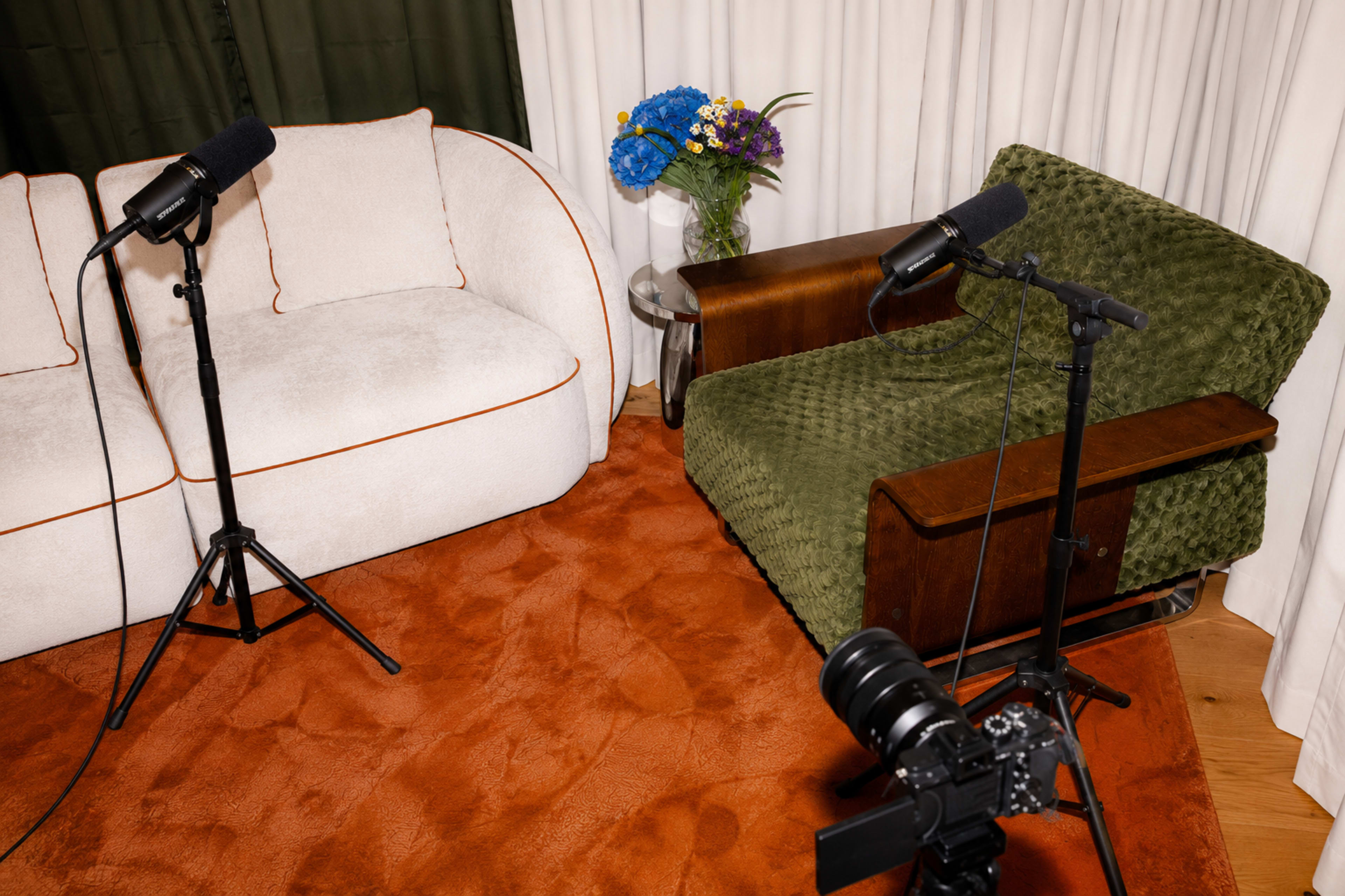 The image shows a room with a light-colored sofa, a green upholstered chair, two microphones on stands, a camera on a tripod, and a vase with flowers on a side table.