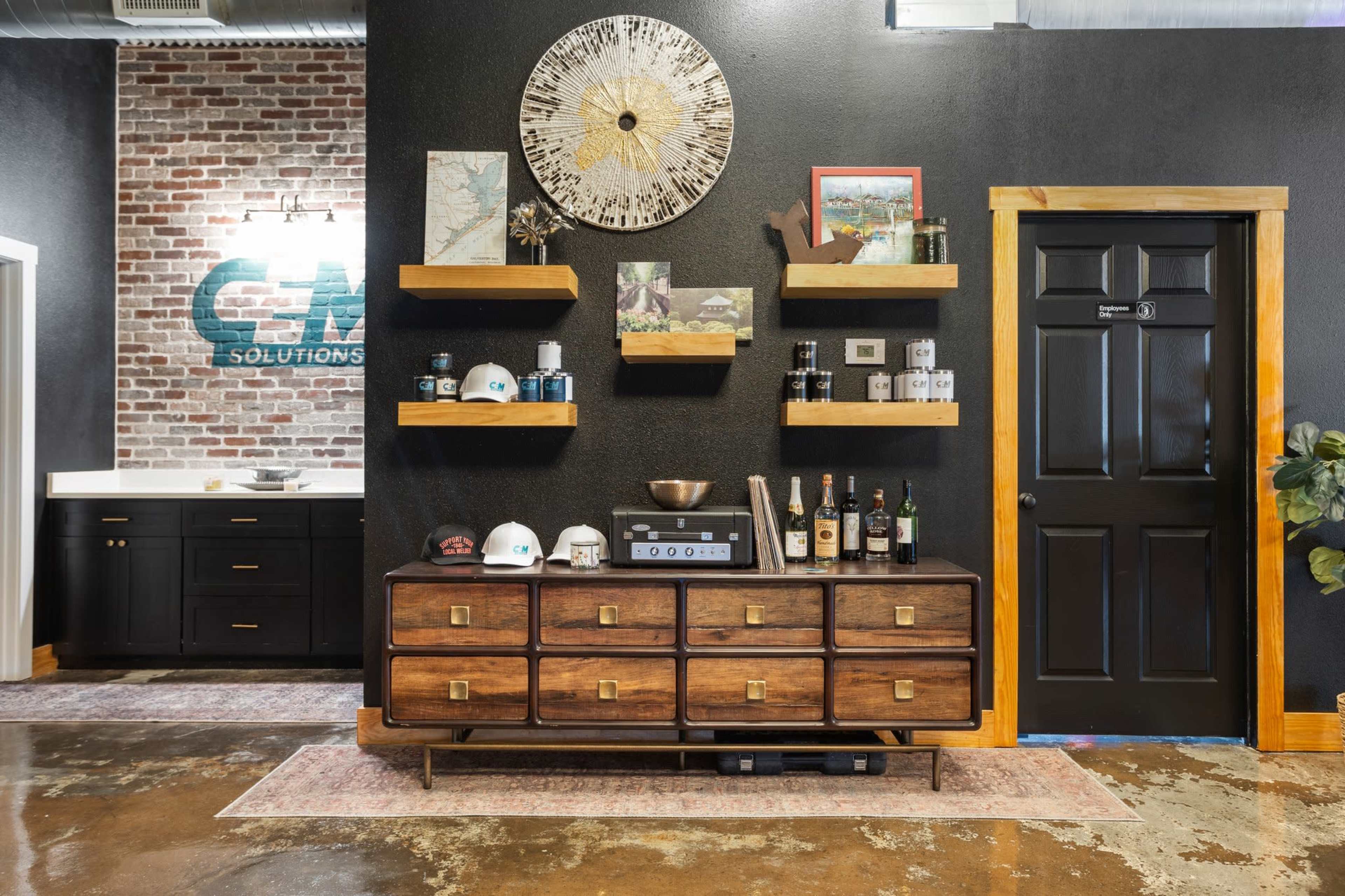 A modern office space featuring a wooden dresser with various items, including bottles and decorative objects, against a black wall with floating shelves and a door leading to another room.