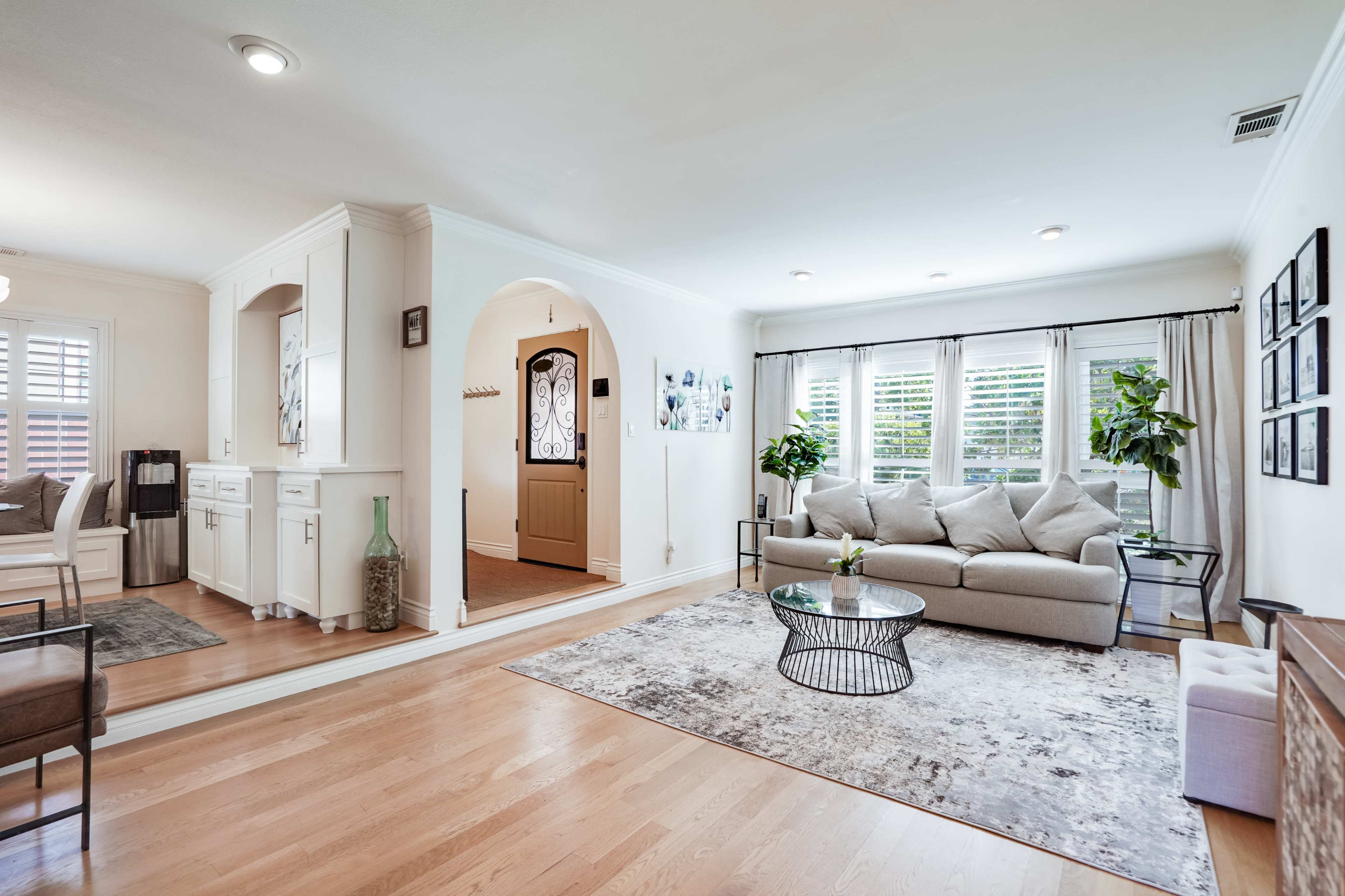A bright, open living room with a couch, coffee table, and large windows, connected to an entrance area featuring a front door and a small kitchenette.