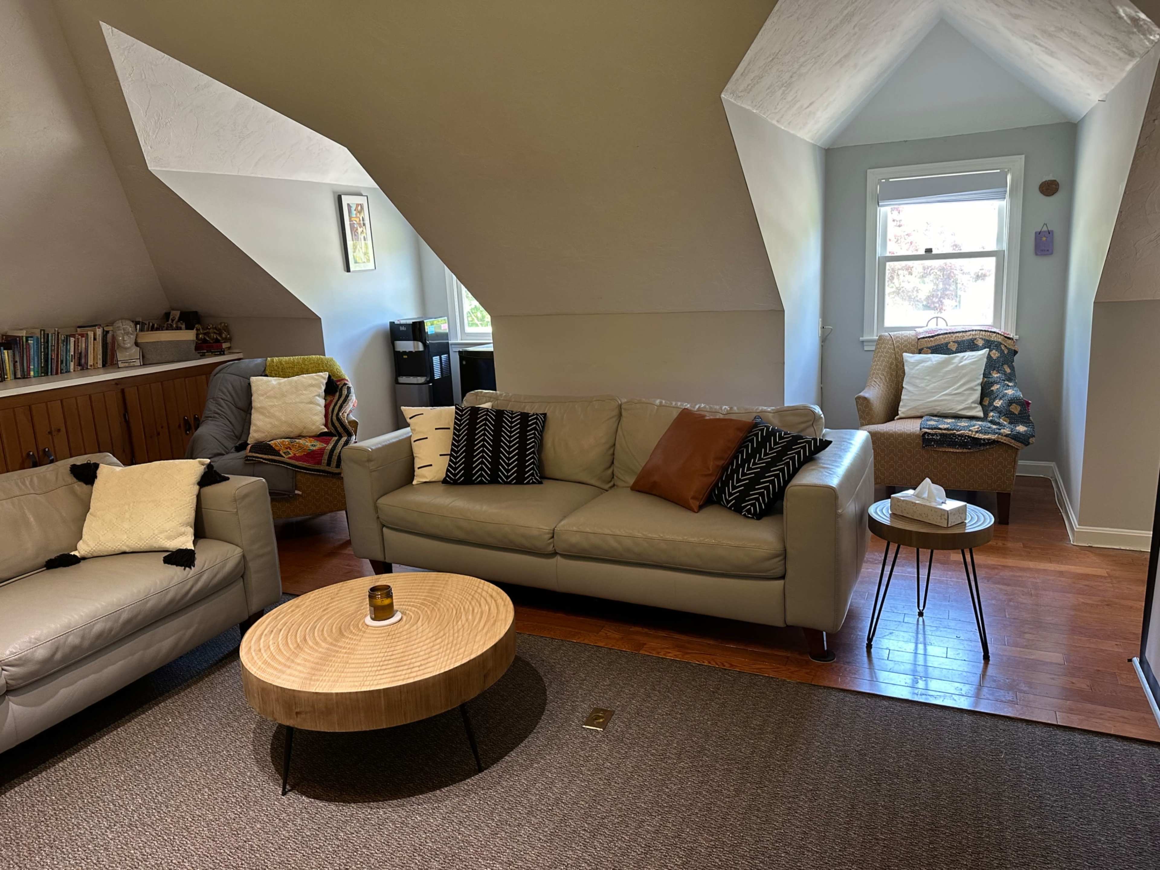 A cozy living room in an attic with a sectional couch, a round wooden coffee table, and a chair by the window.