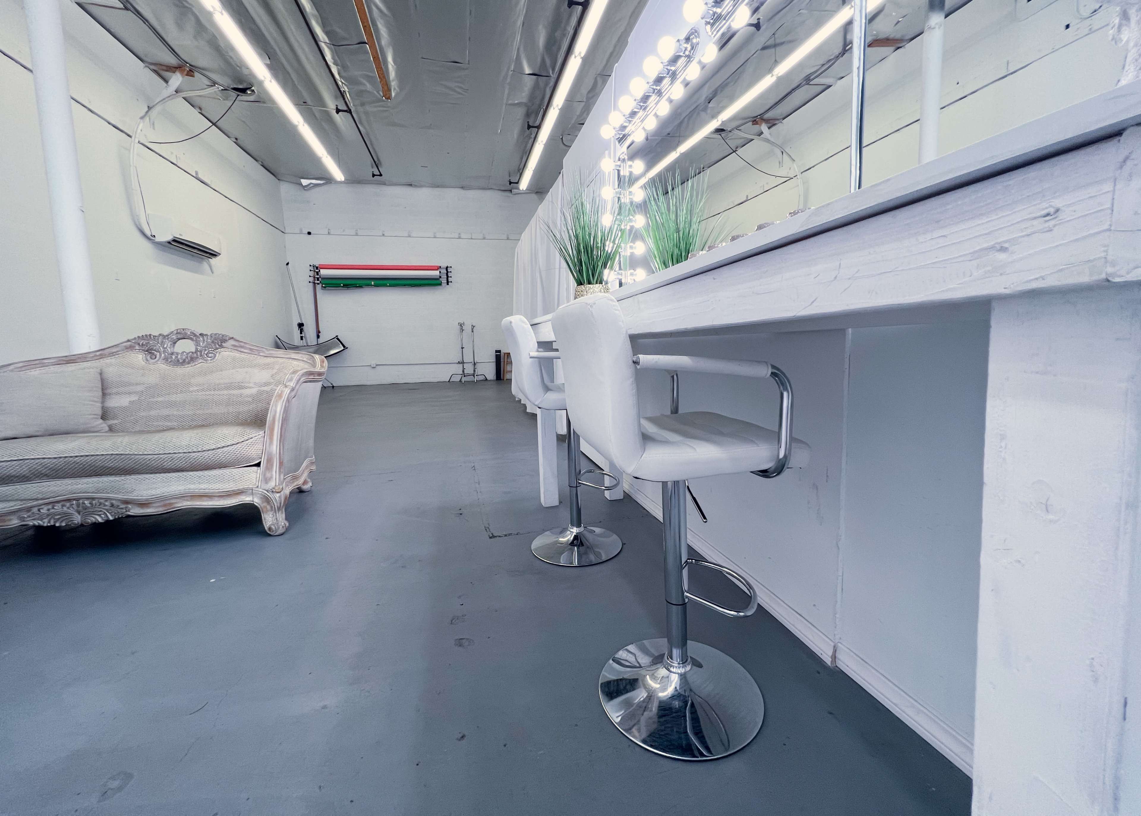 The image depicts a modern vanity area with a row of mirrored lights, white bar stools, and a stylish couch in an empty room.
