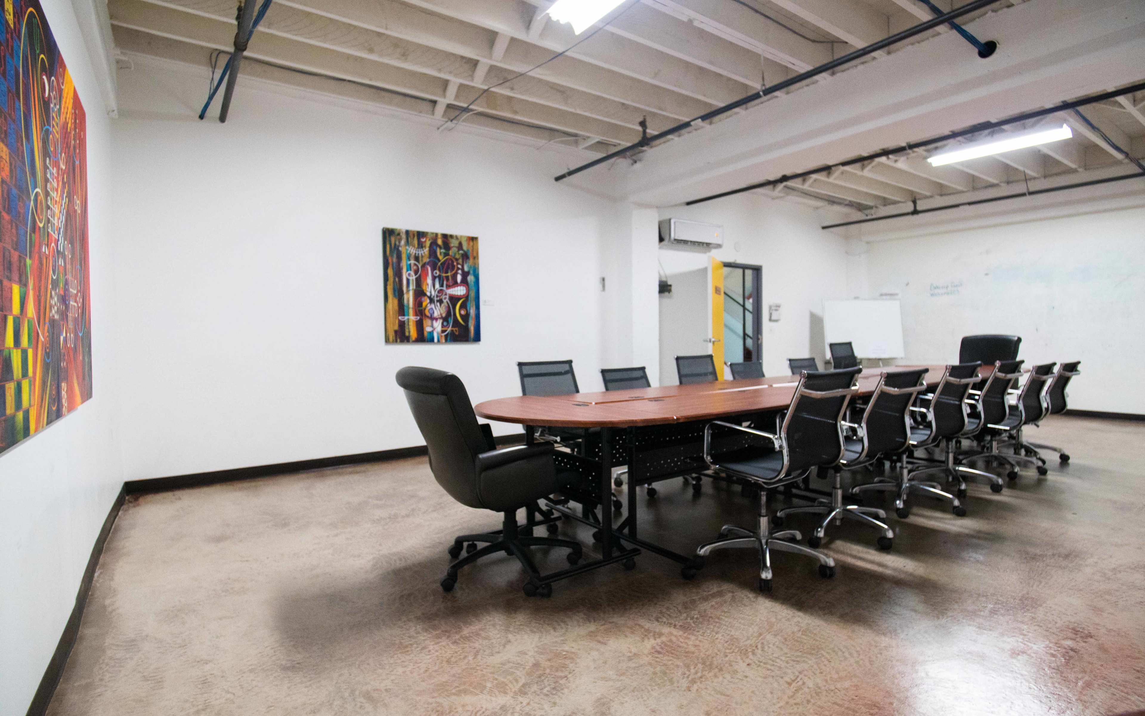 A conference room with a large rectangular table surrounded by office chairs, flanked by two abstract paintings on the walls.