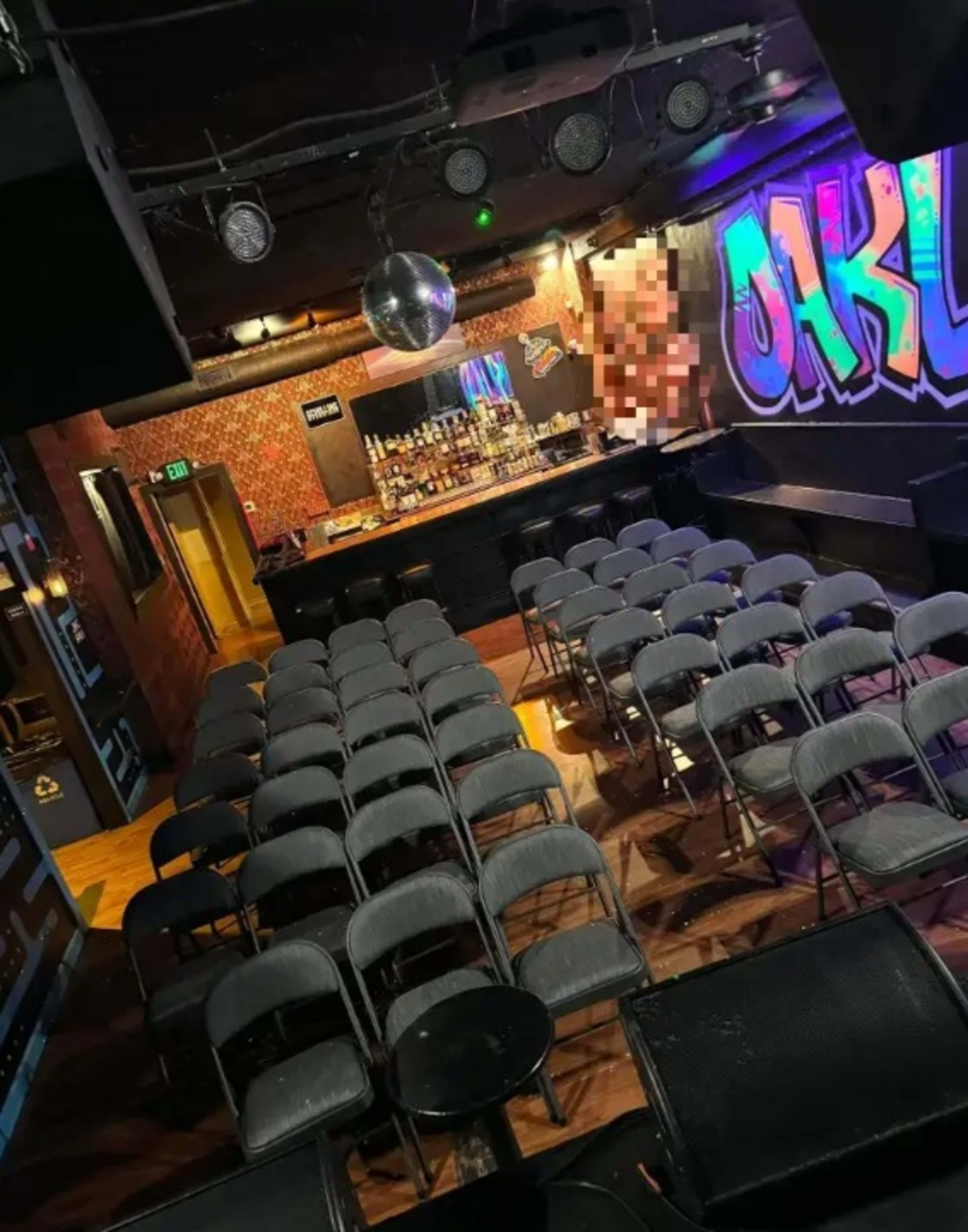 The image shows a seated area in a performance venue with rows of black folding chairs facing a bar counter and colorful graffiti on the wall.