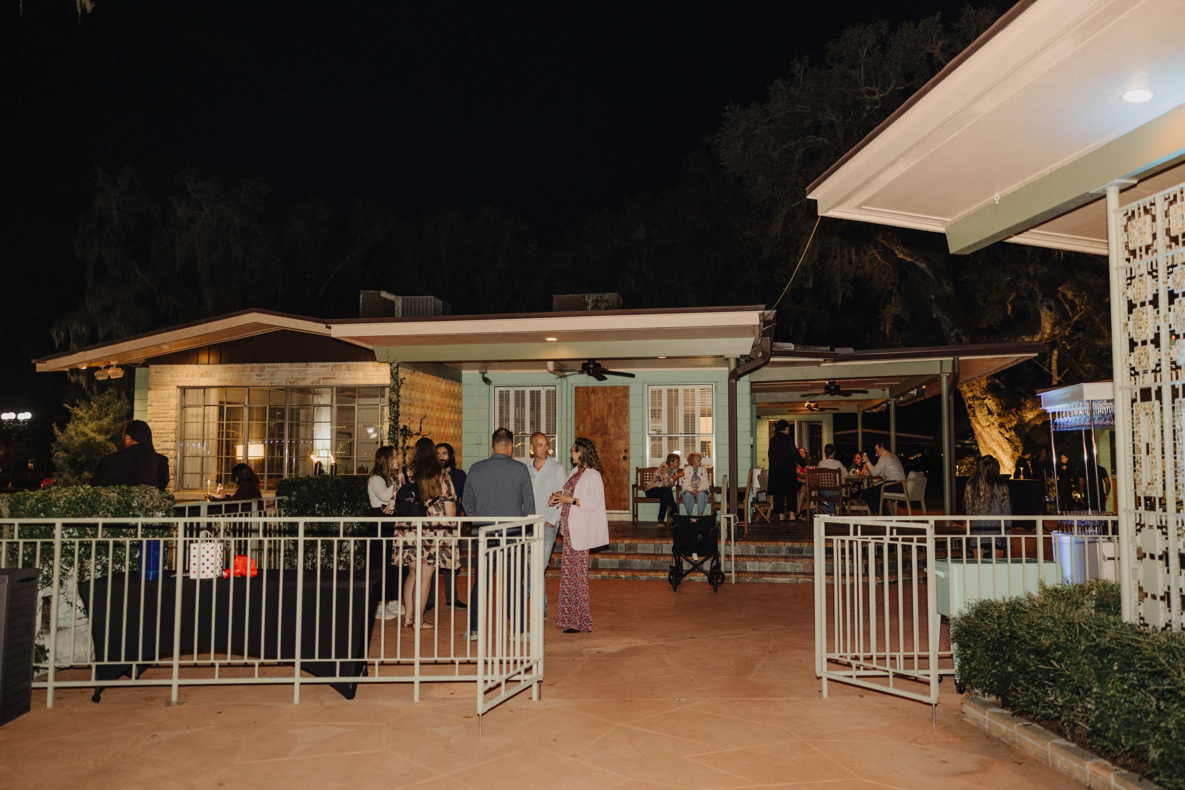 A group of people gather outside a house during the night, with some socializing near a fenced area while others are seated at tables.