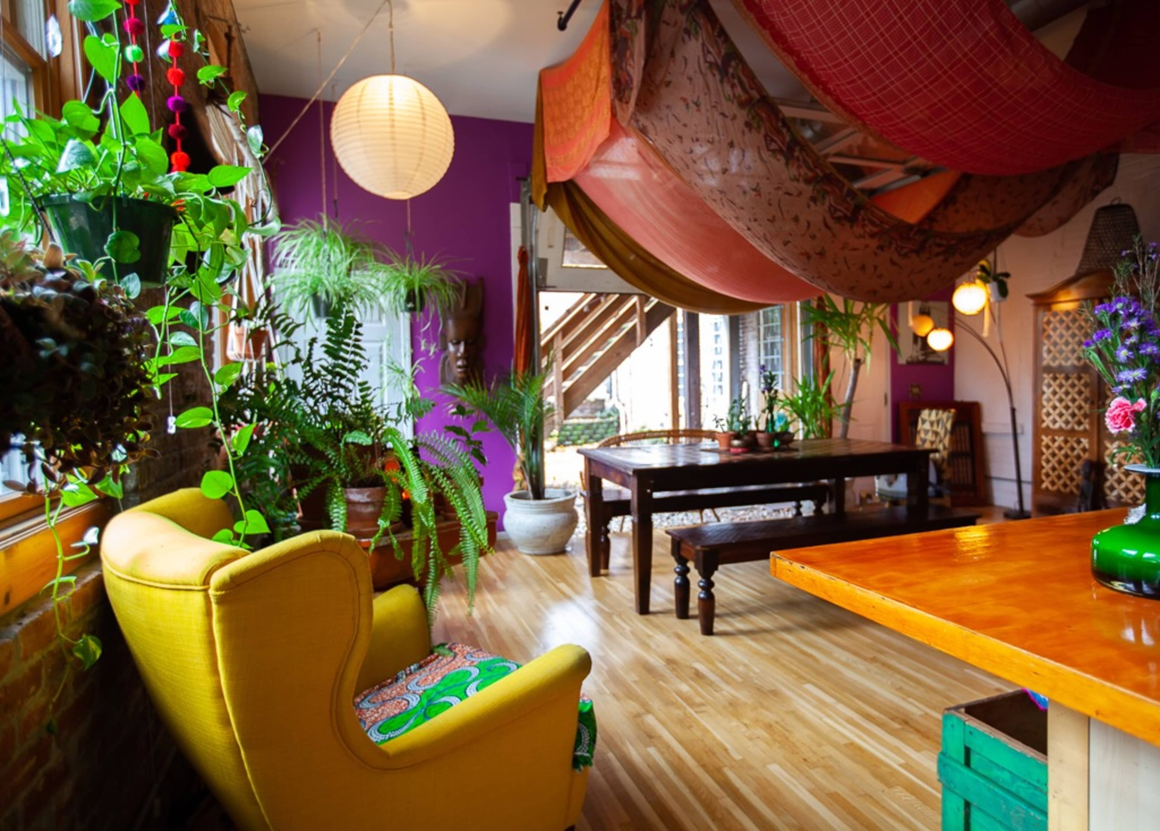 The room features a bright yellow armchair, a large wooden table, and an array of plants, all under colorful drapes and lanterns.
