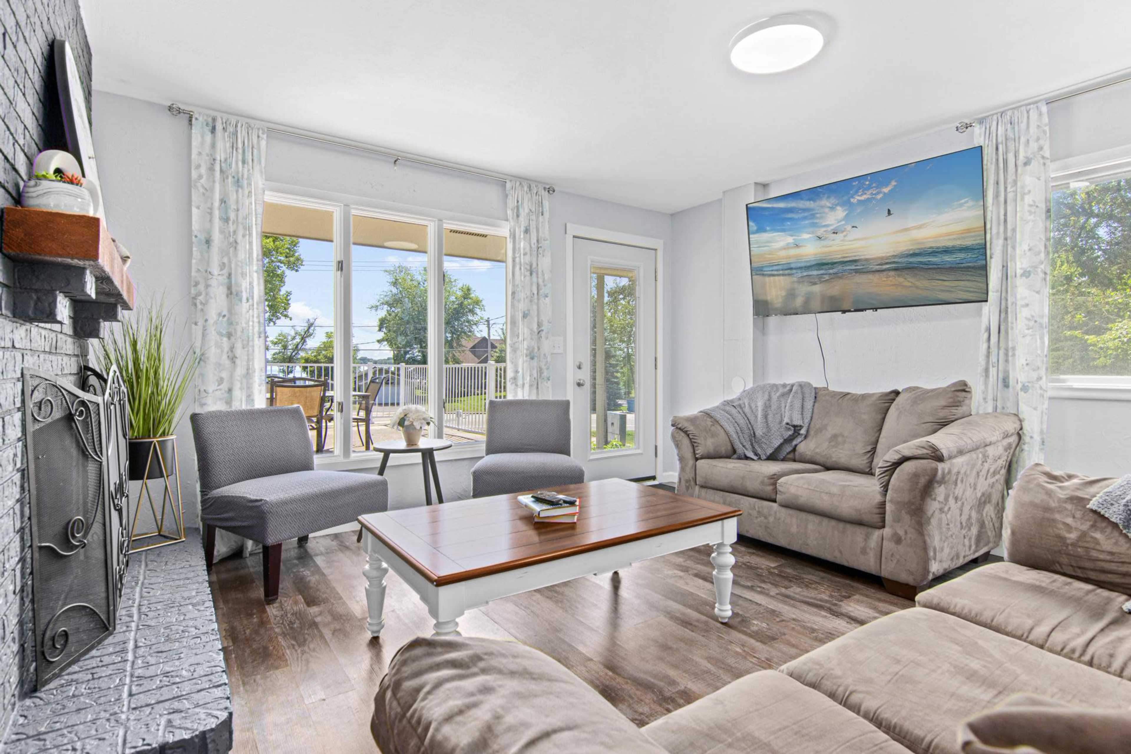 A cozy living room features a large sectional sofa, two armchairs, a wooden coffee table, and a mounted television, with large windows letting in natural light.