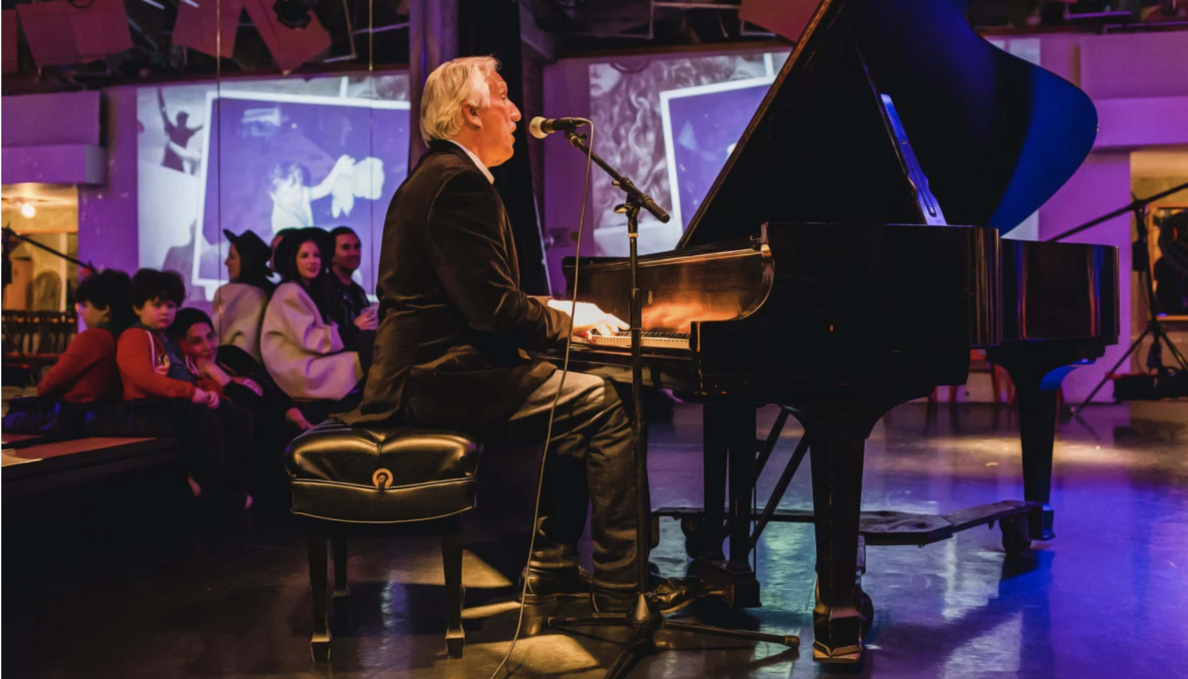 A man sings and plays a grand piano on stage while an audience, including children, watches attentively in a brightly lit venue.