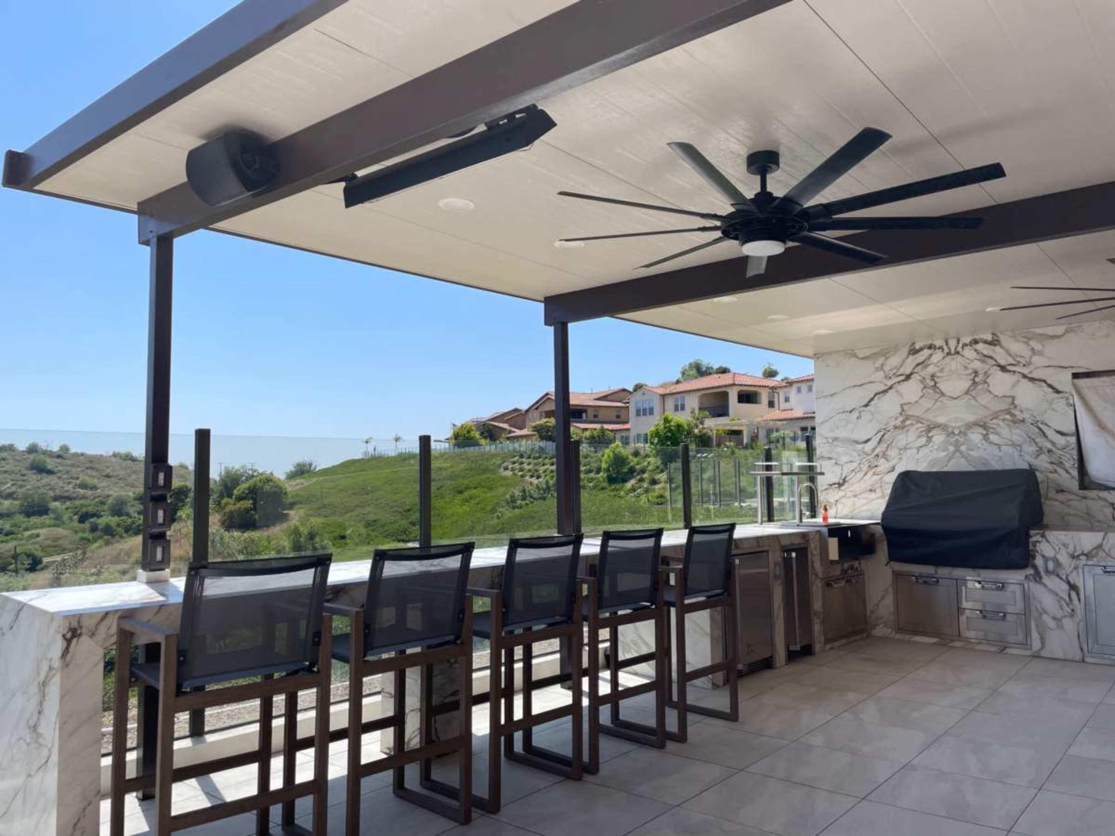 An outdoor kitchen with a marble countertop and bar seating, featuring a built-in grill and a ceiling fan, overlooking a green hillside and homes in the background.