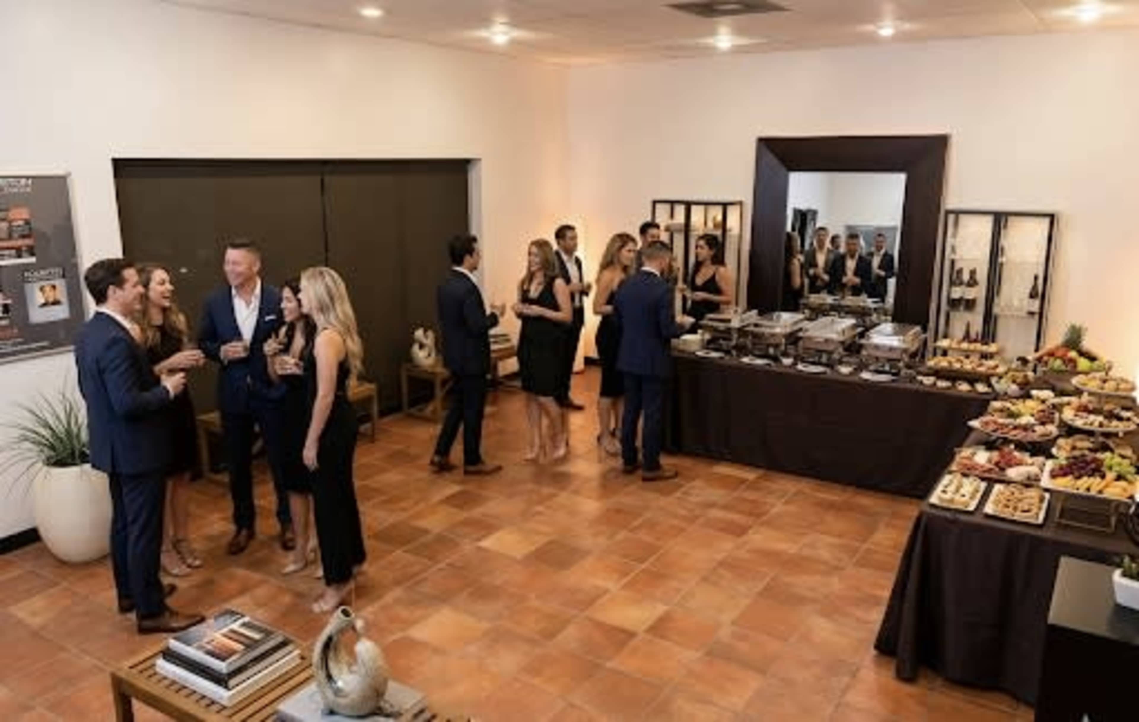 A group of people in formal attire socialize in a warm, well-lit room while a buffet of food is displayed on a table nearby.