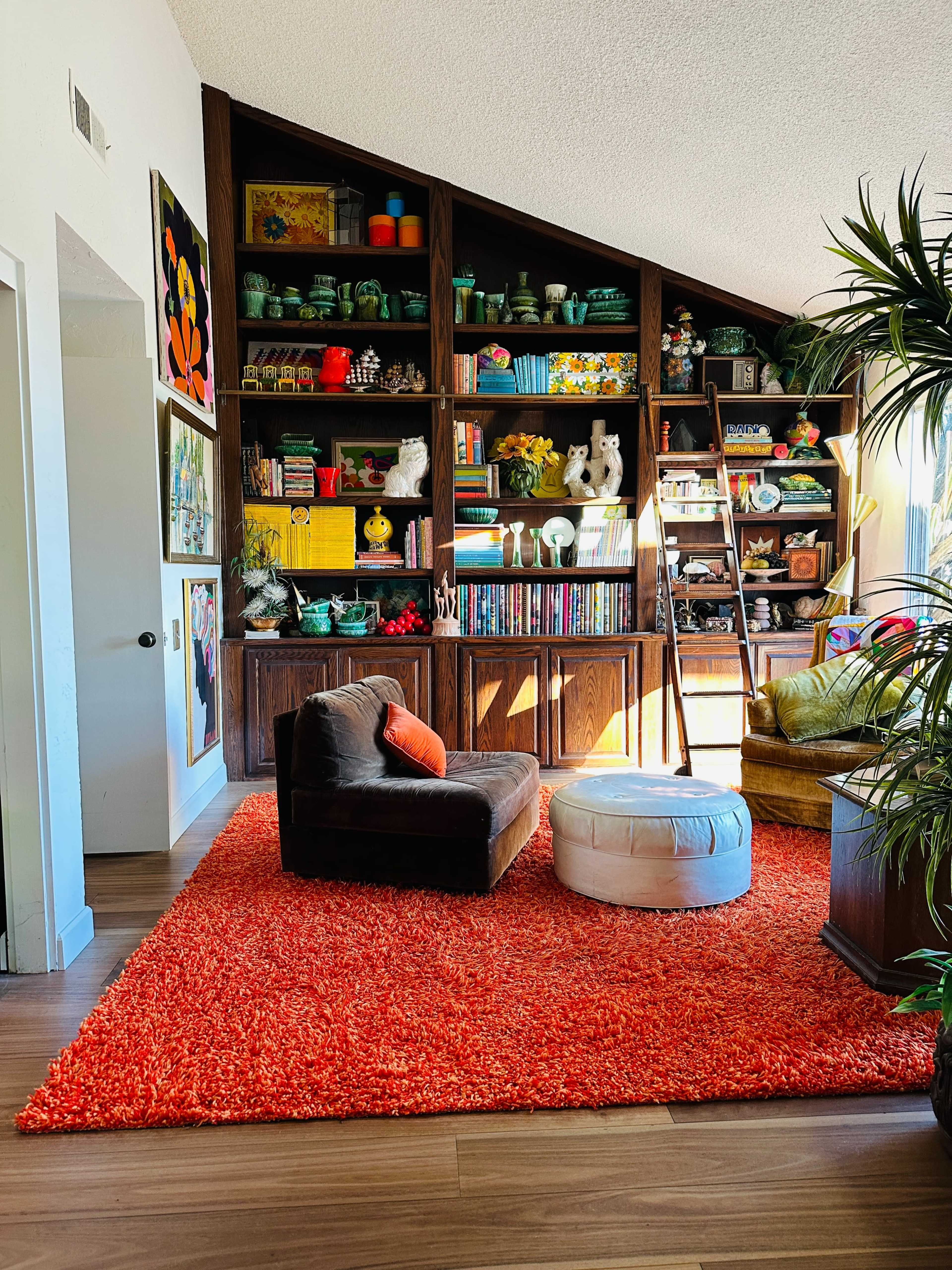 The image shows a cozy living room with a brown armchair, a white ottoman, and a vibrant orange shag rug, accompanied by a wooden bookshelf filled with colorful decorative items and plants.