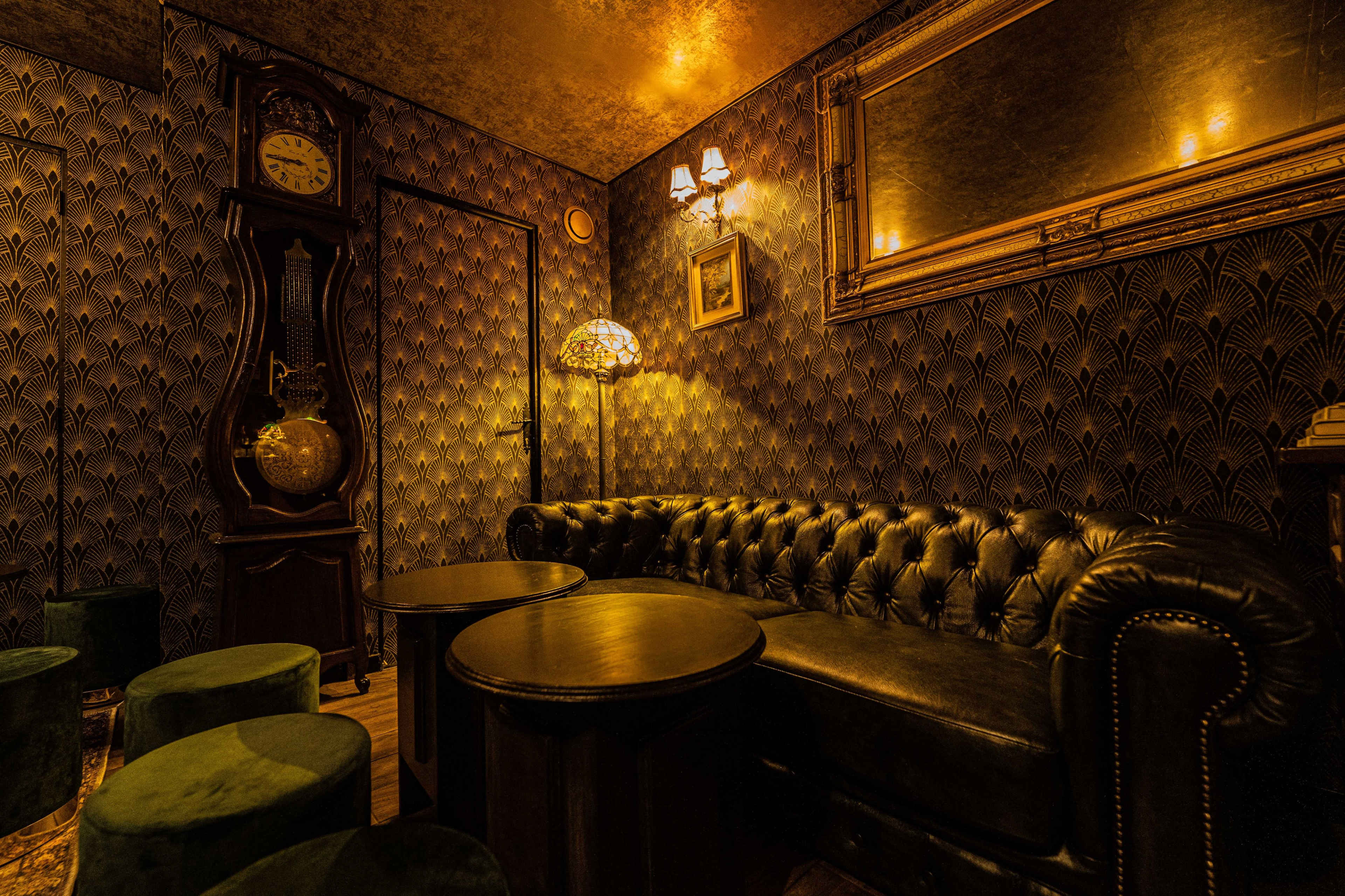 A dimly lit room with dark, patterned walls, a leather chesterfield sofa, and small round tables, featuring a vintage clock and decorative lamp.