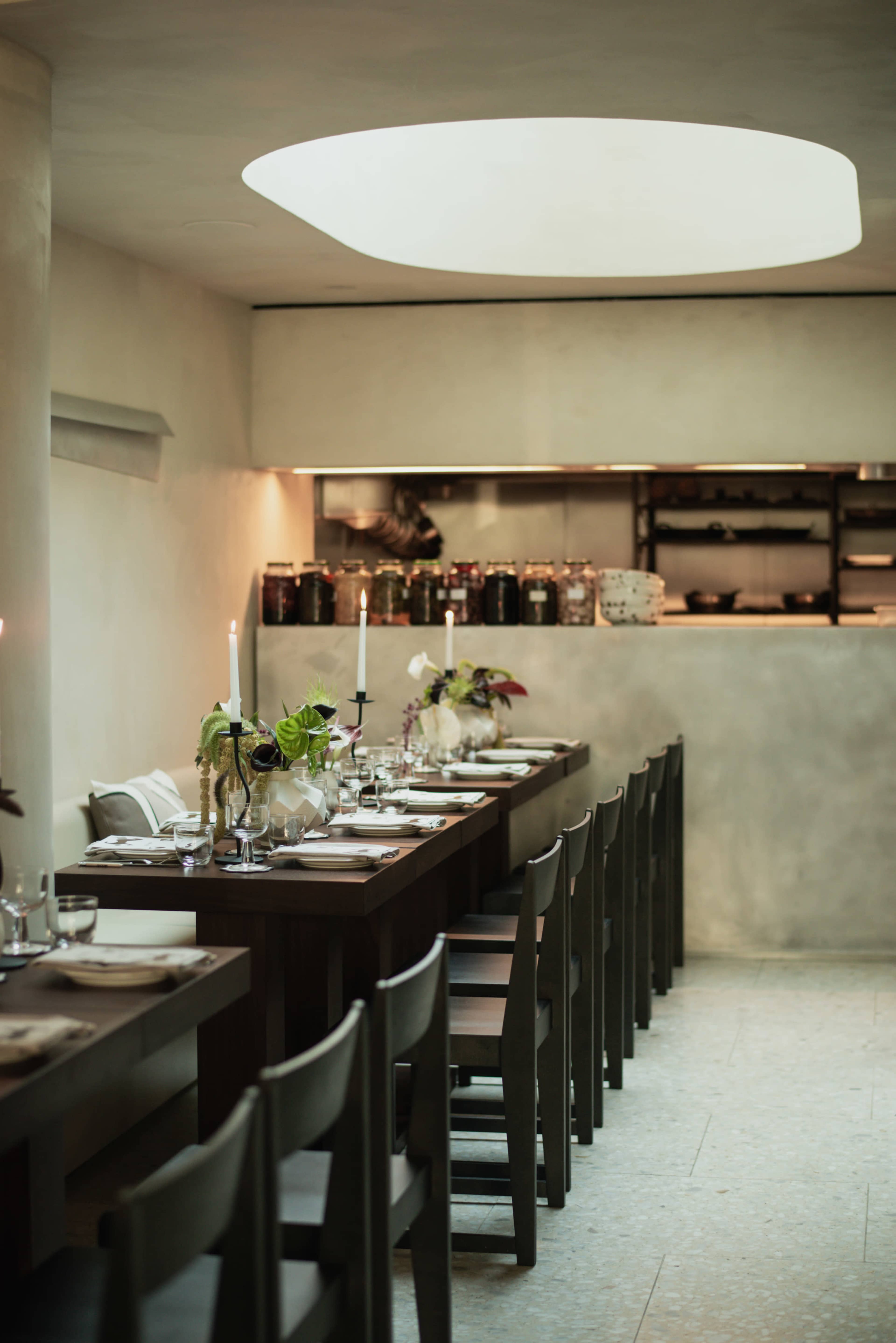 A long, wooden dining table is set with candles and glassware in a minimalist restaurant with soft, natural lighting and jars arranged on shelves in the background.