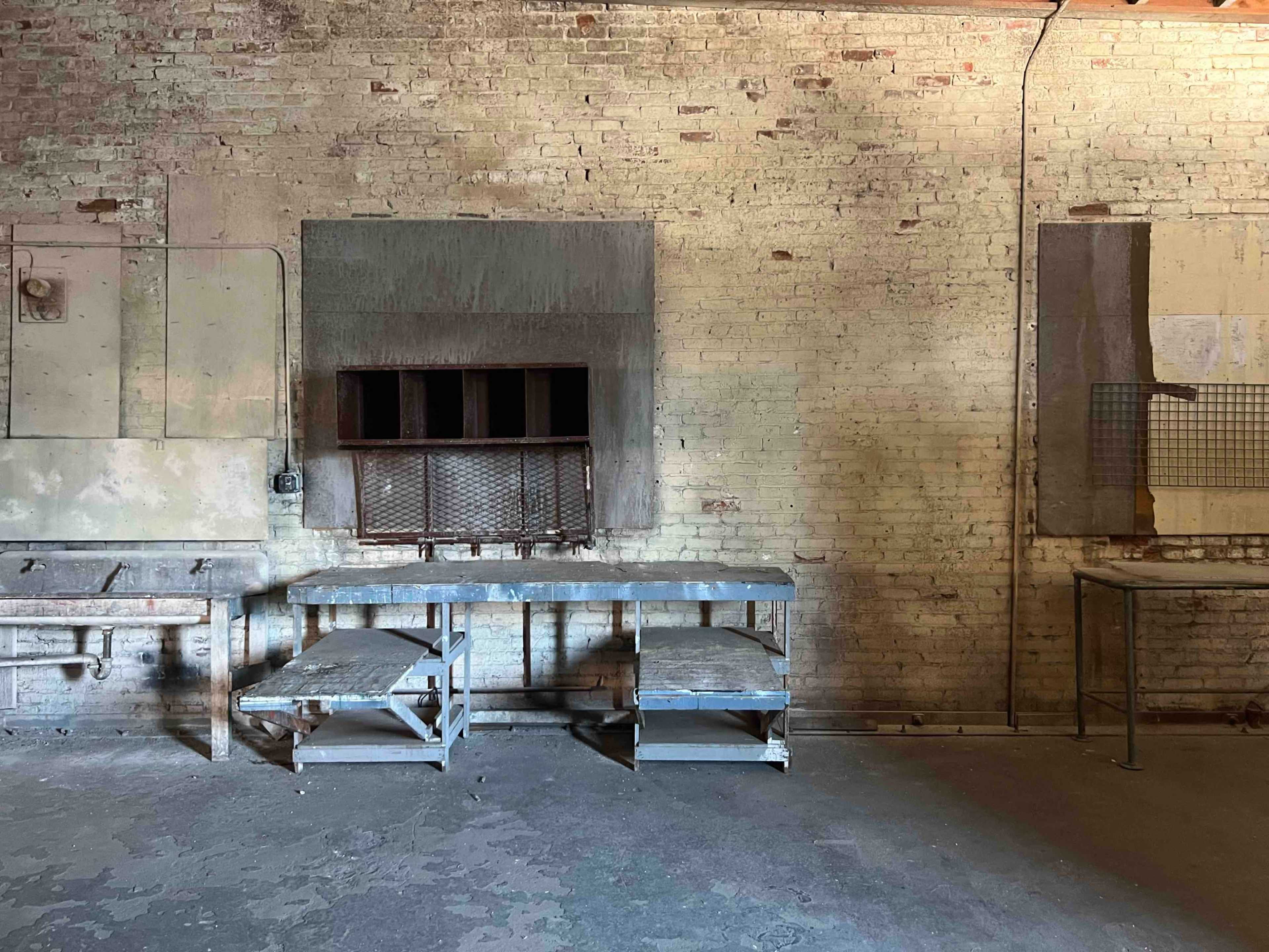 An empty industrial space with two metal tables against a brick wall, featuring a large window and various wall fixtures.