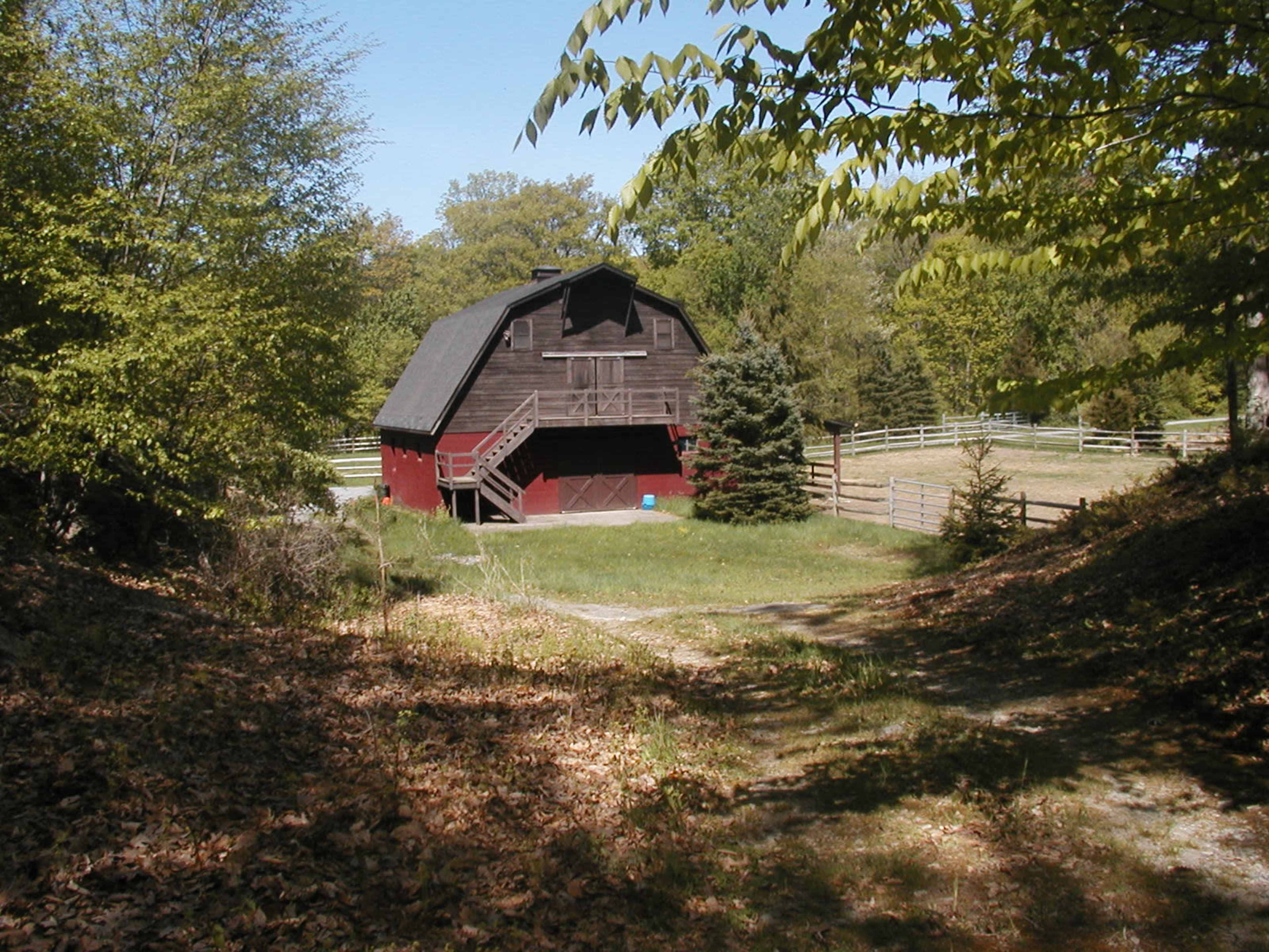 A red barn is situated at the end of a grassy path, surrounded by trees and a wooden fence.
