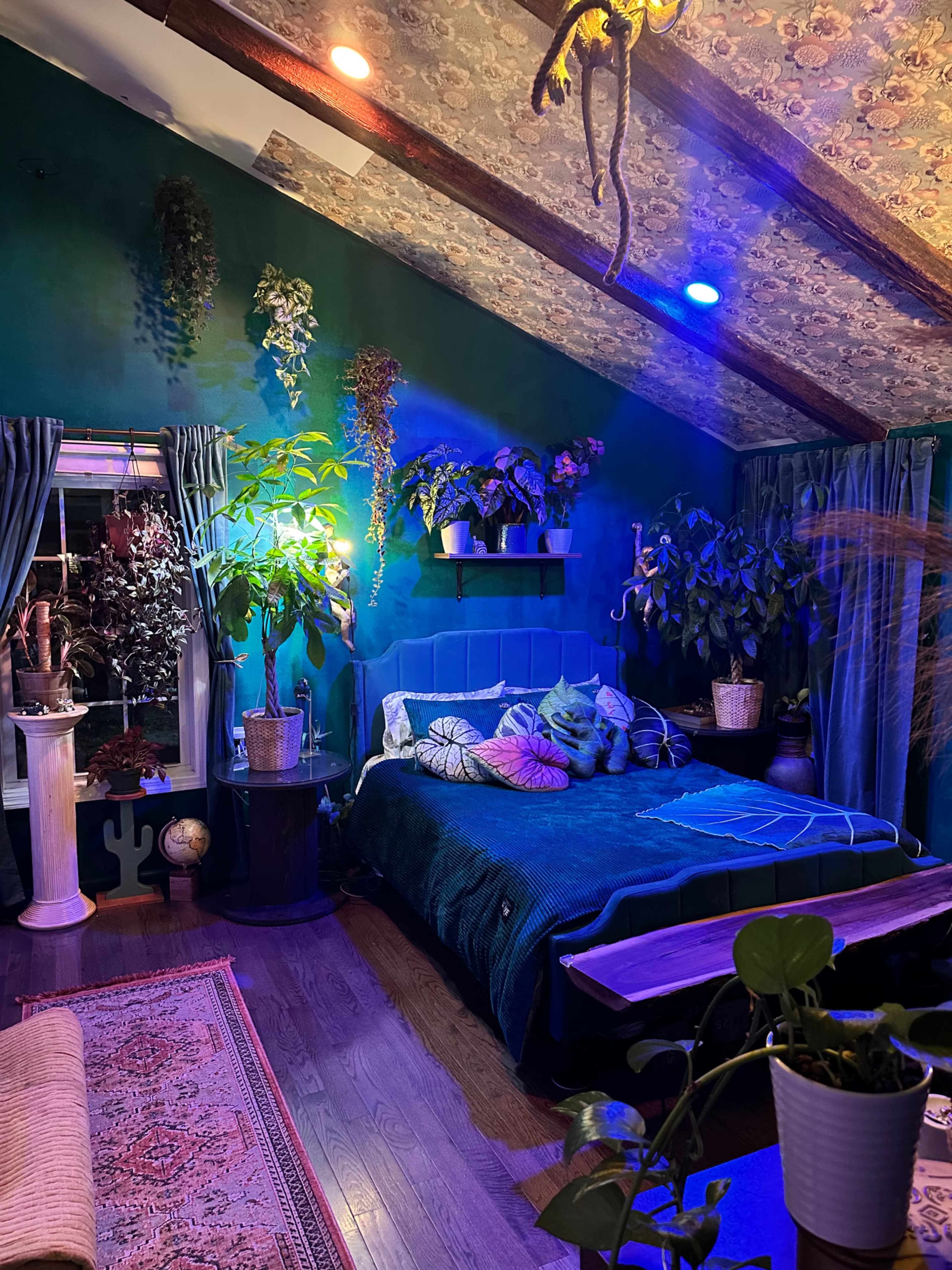 The image shows a cozy bedroom with a green color scheme, featuring a large bed surrounded by various potted plants and illuminated by colorful lights.