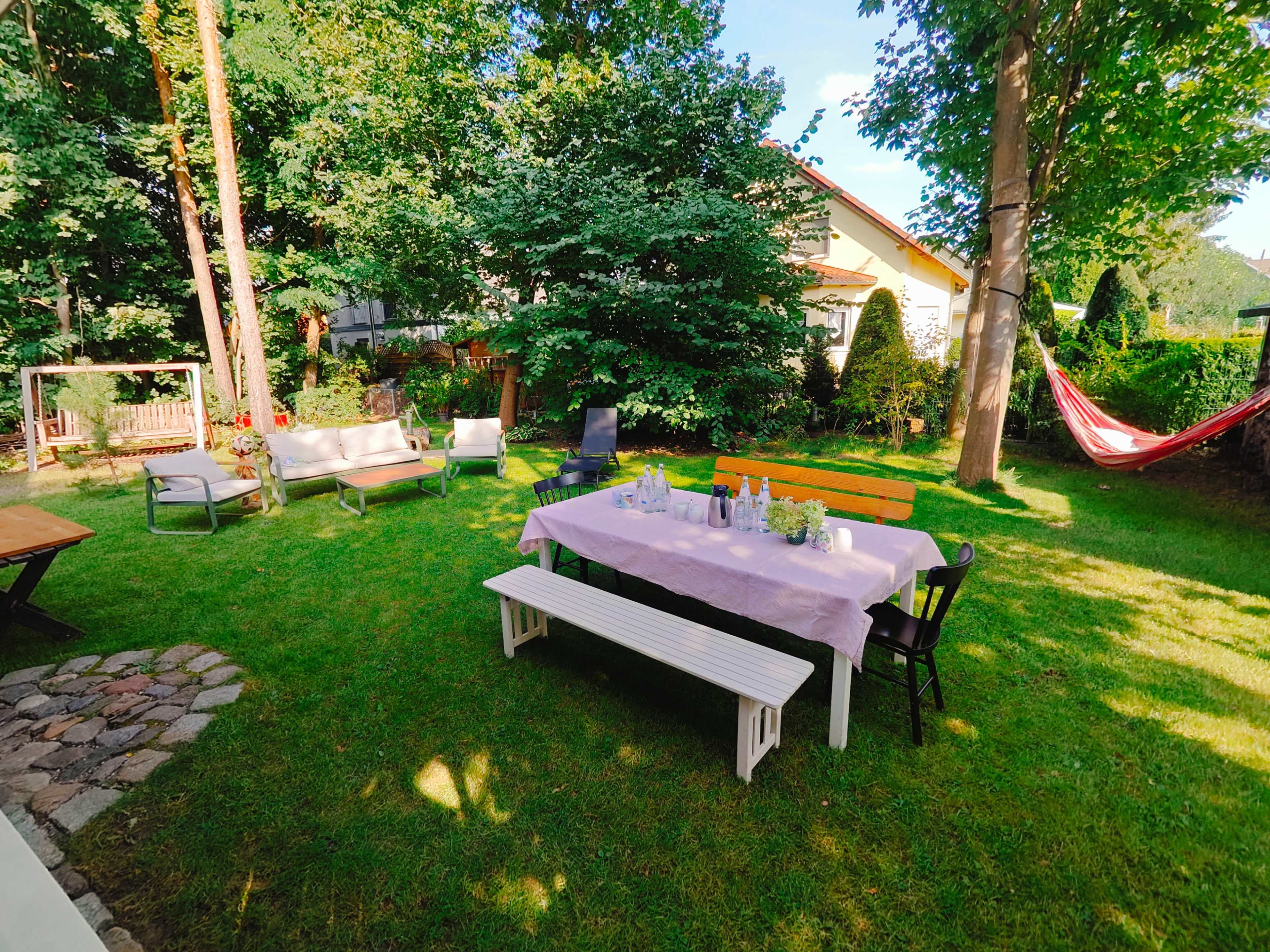 A neatly arranged garden features a table with fresh flowers, surrounded by chairs, benches, and a hammock, all set against a backdrop of lush greenery.