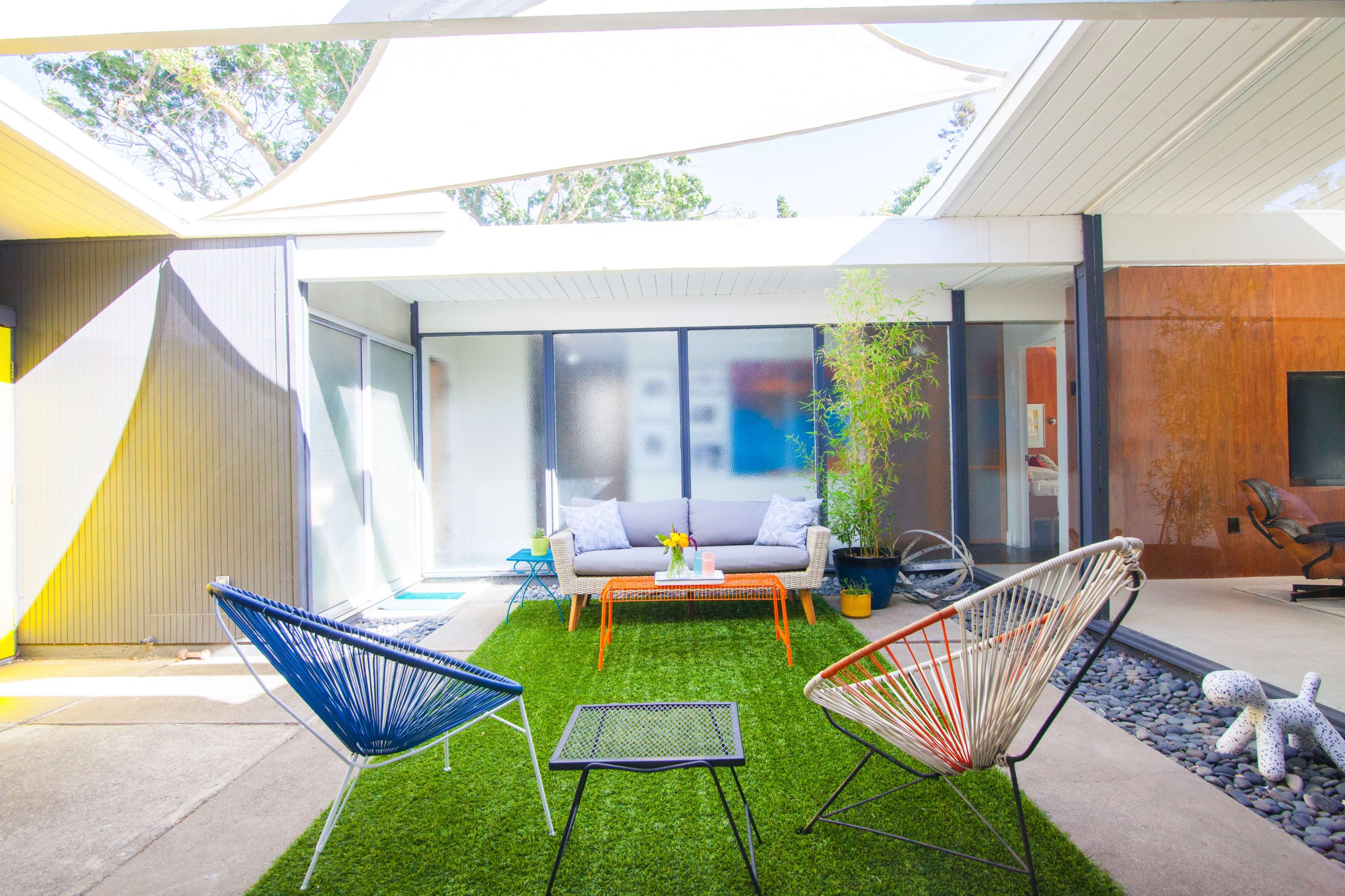 A bright outdoor living space features colorful chairs, a low table, and greenery, surrounded by modern glass walls.