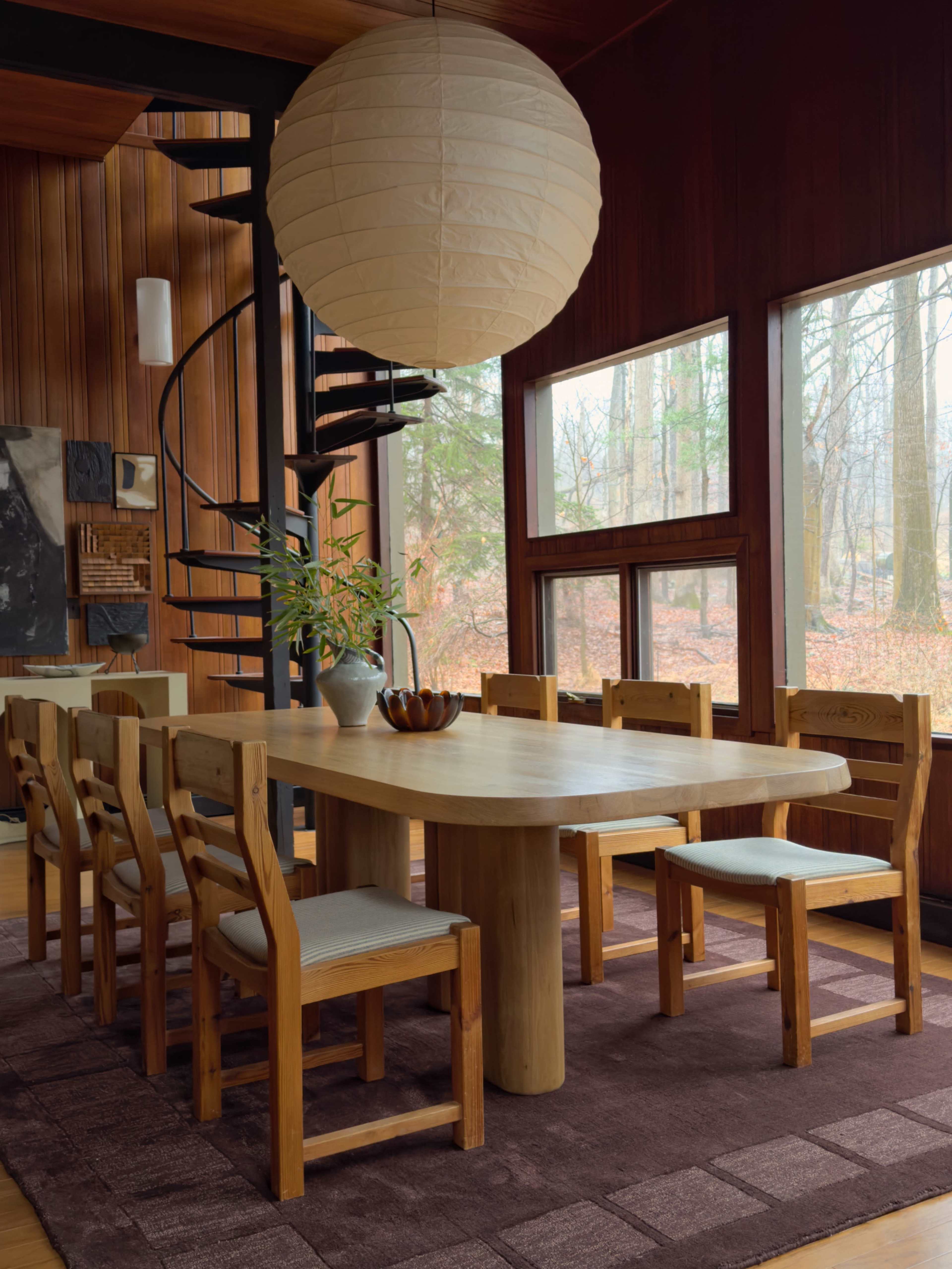The image shows a wooden dining table surrounded by eight chairs, situated in a room with large windows that offer a view of a wooded area, complemented by a spiral staircase and a round pendant light overhead.