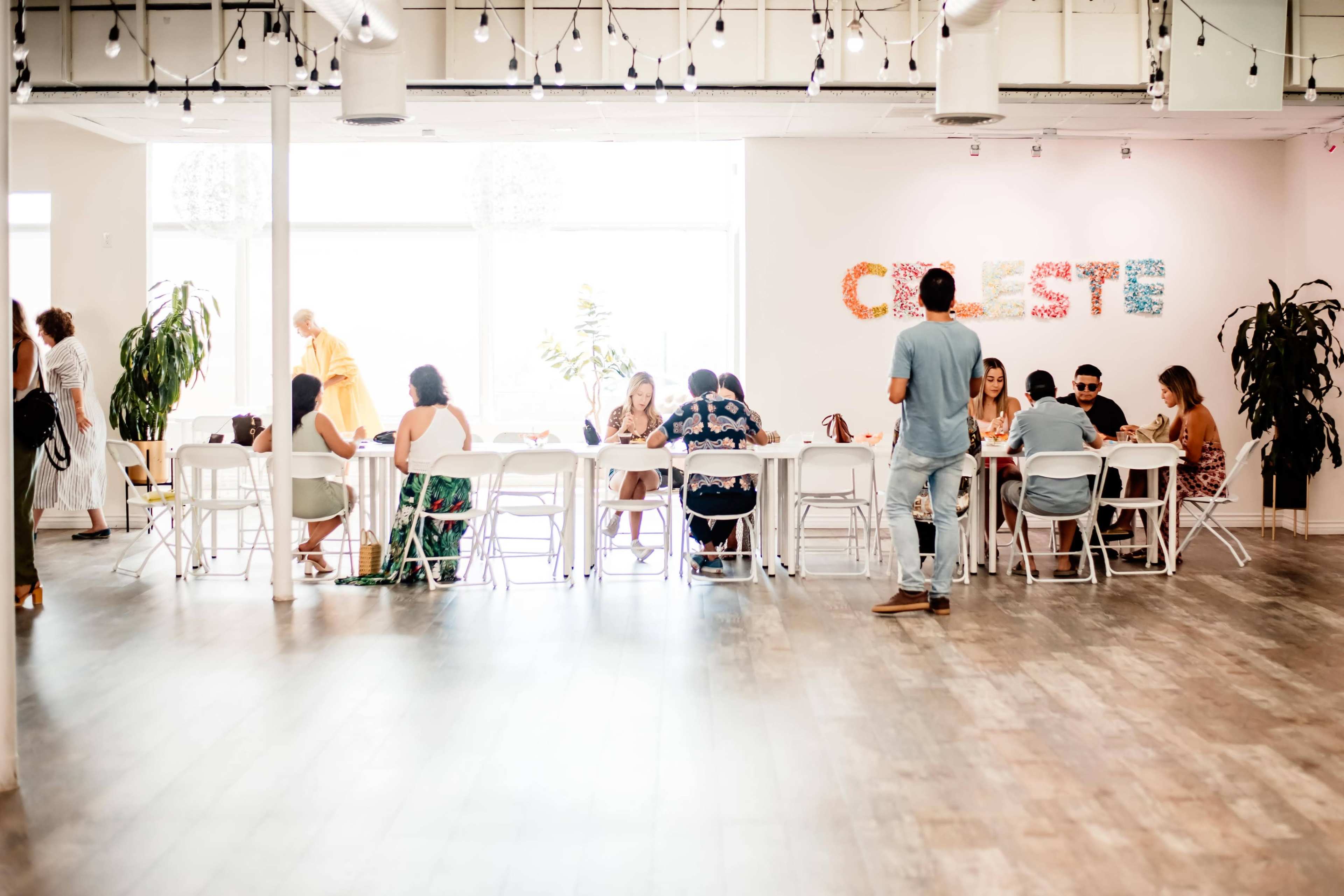 A group of people sits at long tables in a brightly lit room decorated with colorful letters spelling "CELESTE" on the wall.
