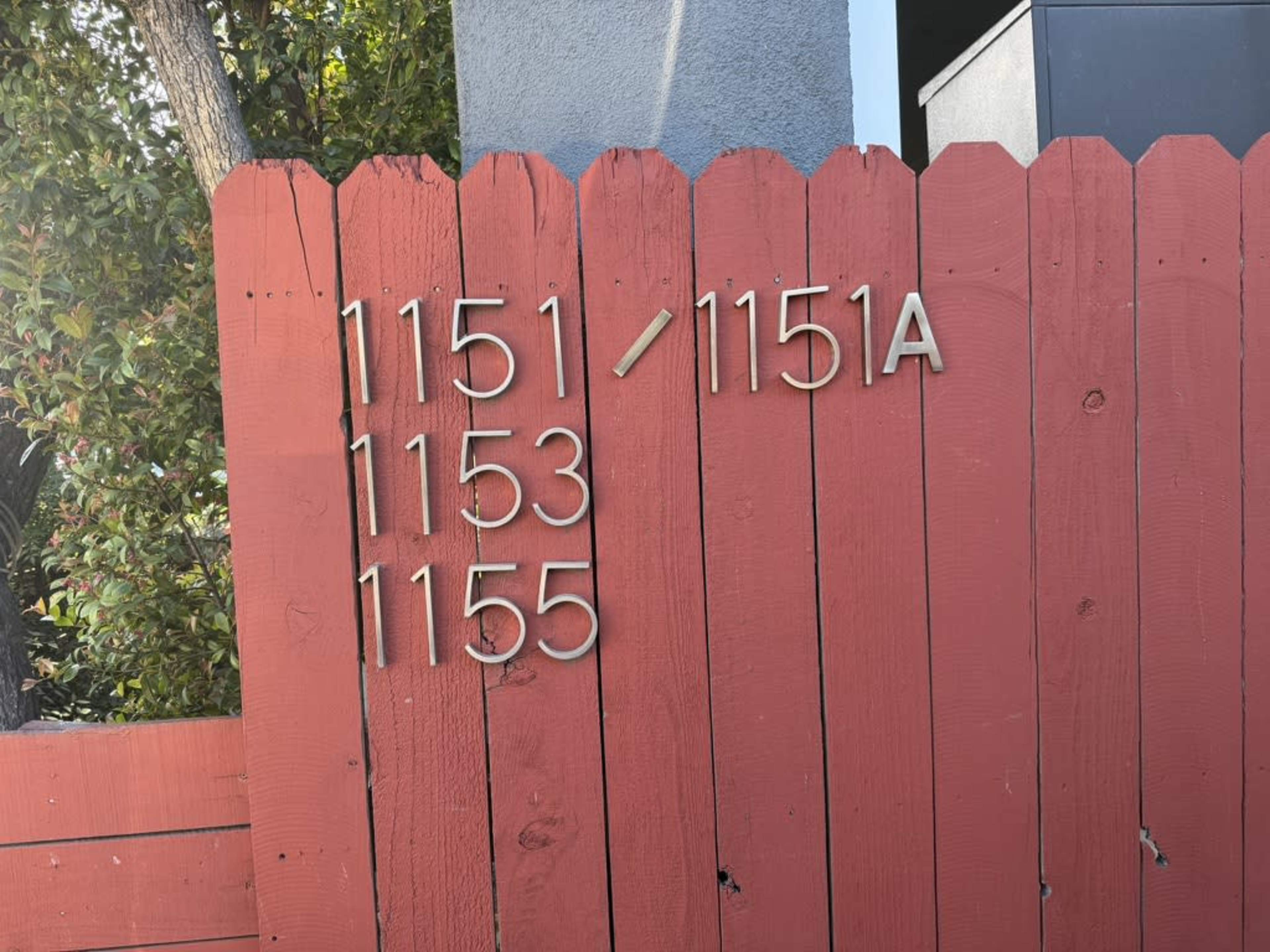 The image shows a red wooden fence with house numbers 1151, 1153, and 1155 displayed in metal numerals, along with 1151A.
