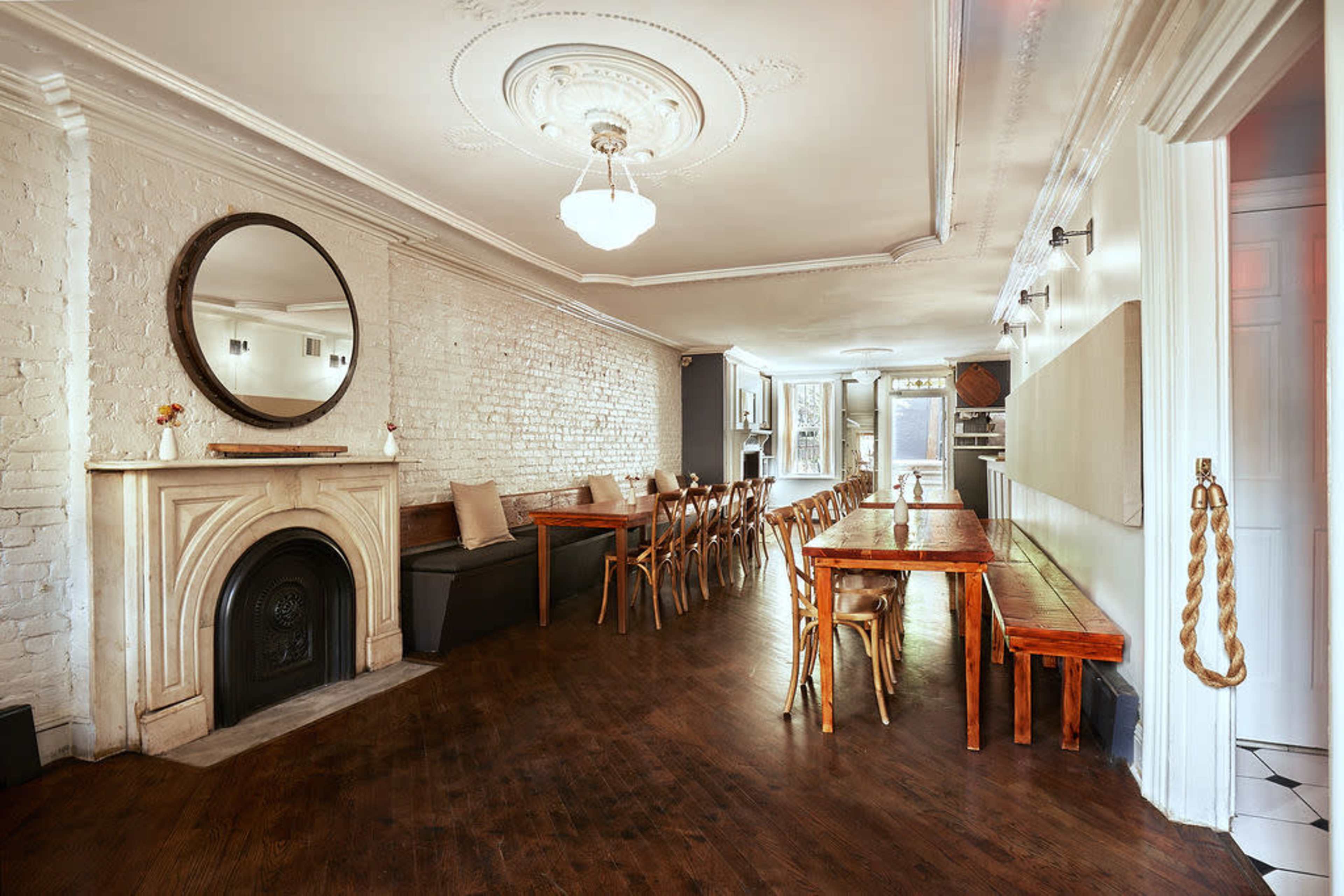 The image shows a spacious room with wooden tables and chairs arranged along brick walls, featuring a decorative mirror and a fireplace.
