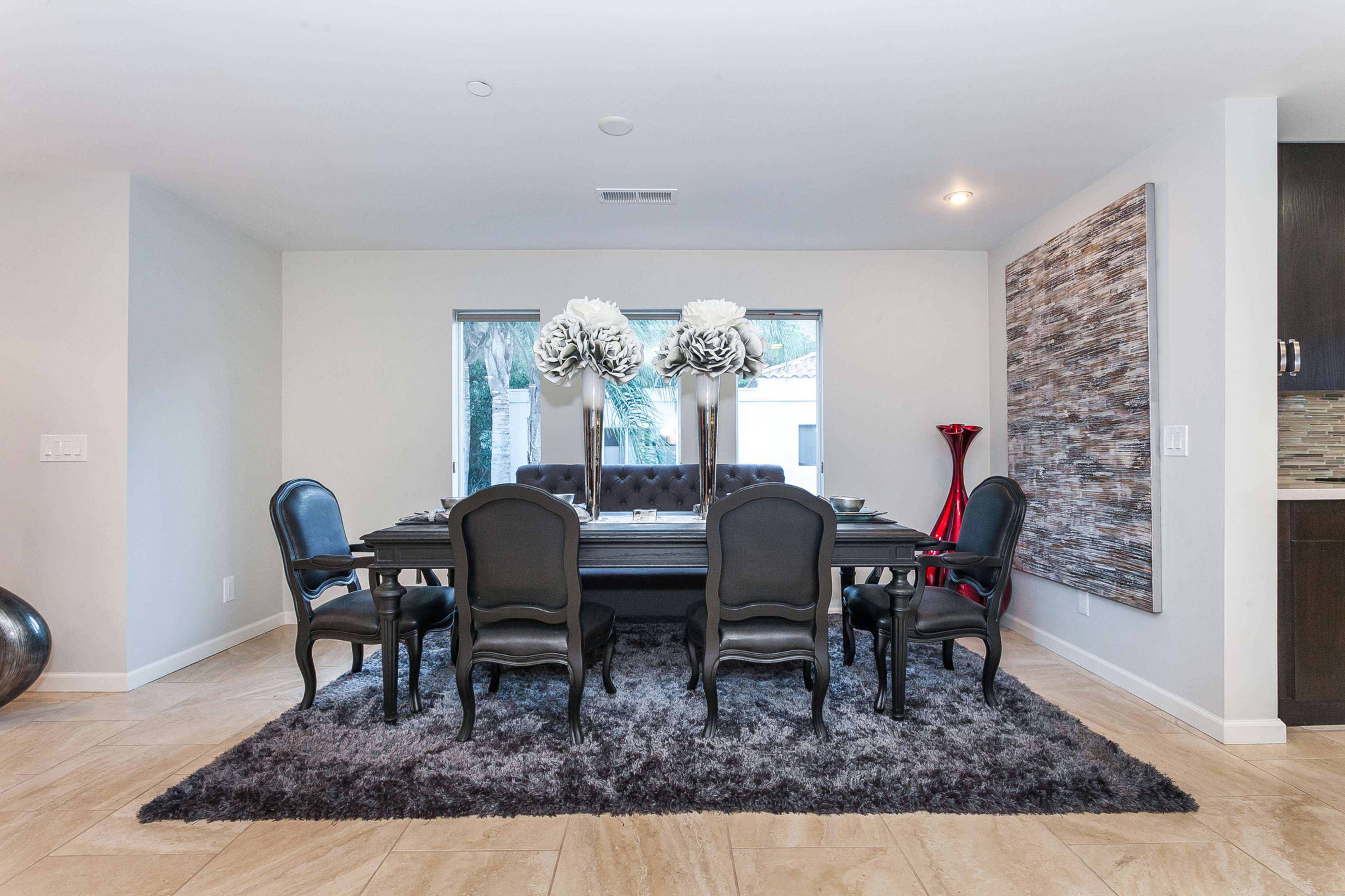 A modern dining room features a large table surrounded by black chairs, with decorative tall flowers at the center and a plush rug underneath.