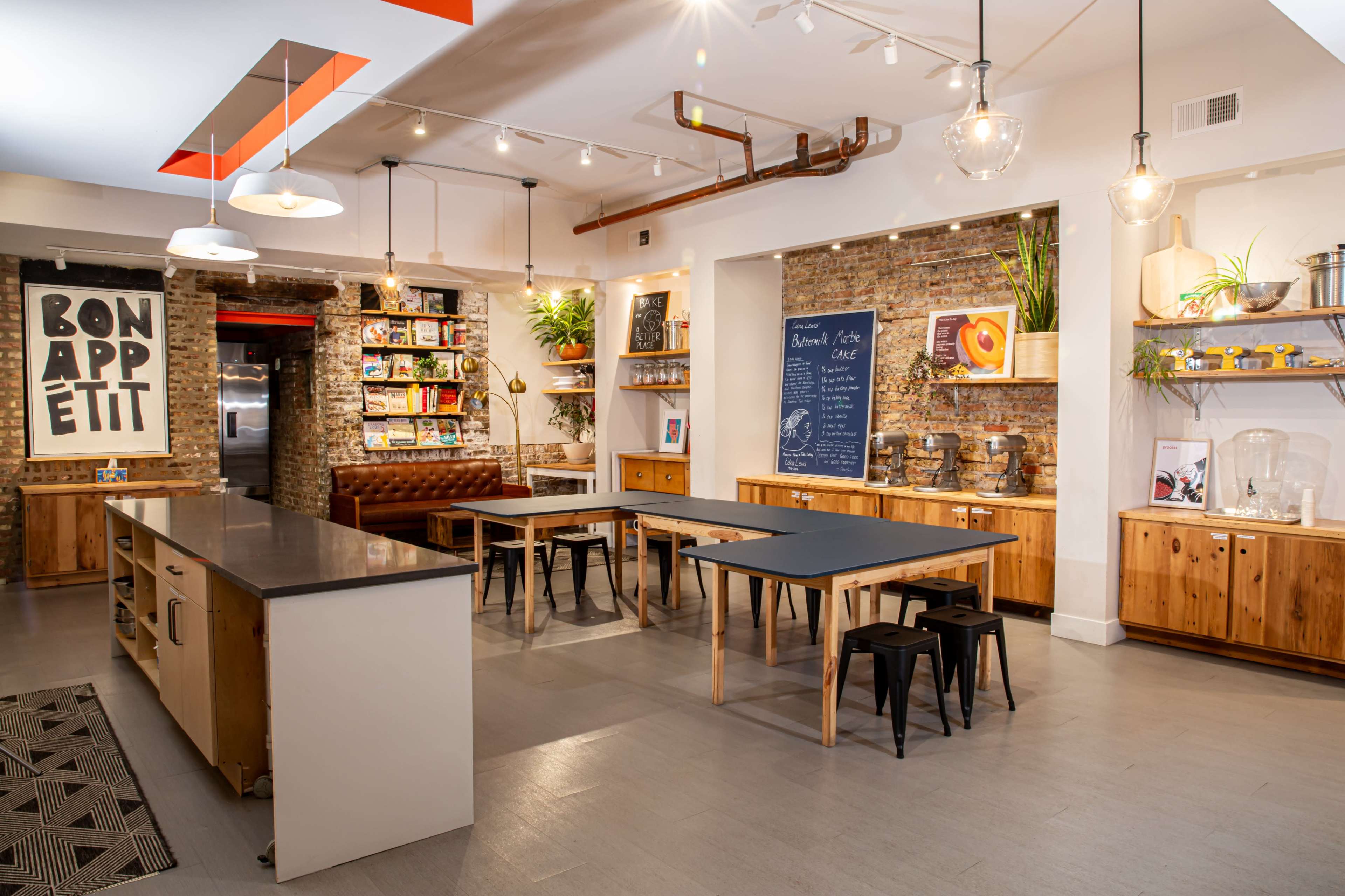 A modern kitchen space features wooden tables, shelves with cookbooks, and a large chalkboard, all set against a brick wall.