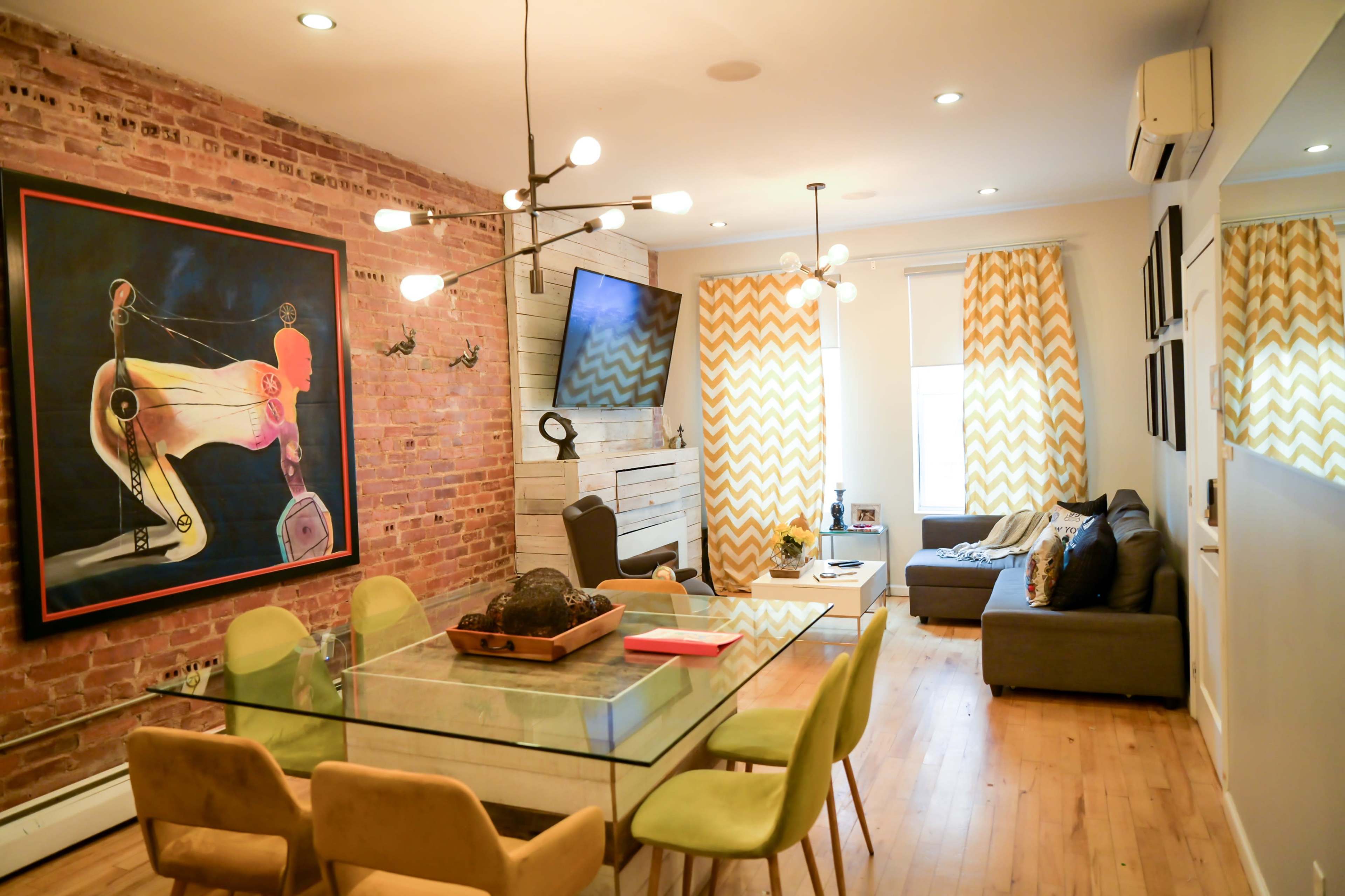 A modern living space with exposed brick walls, a glass dining table surrounded by green chairs, and a seating area featuring a gray sofa and decorative curtains.