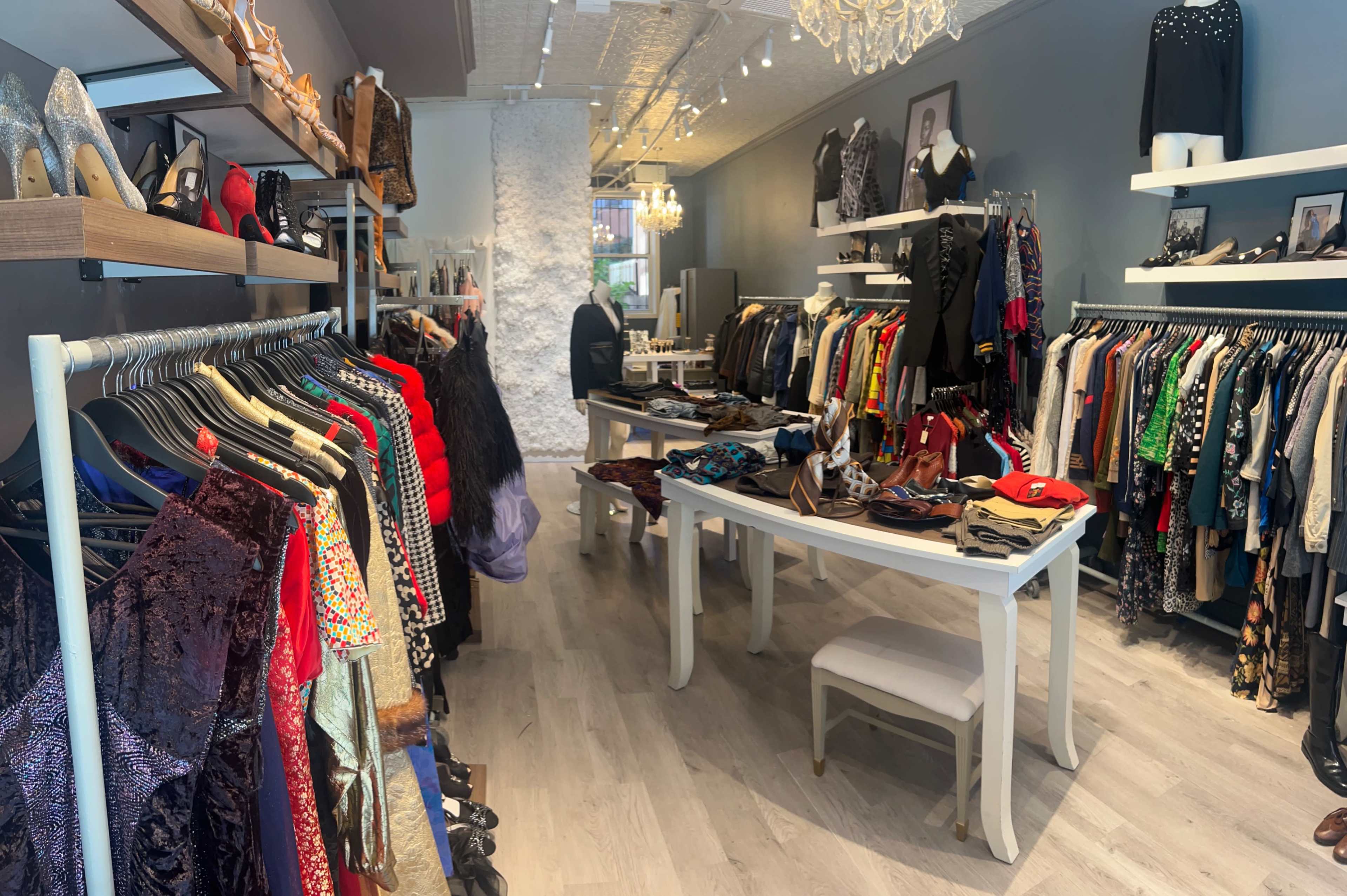 The image shows a boutique clothing store with racks of garments and accessories displayed on tables, featuring a variety of colors and styles.
