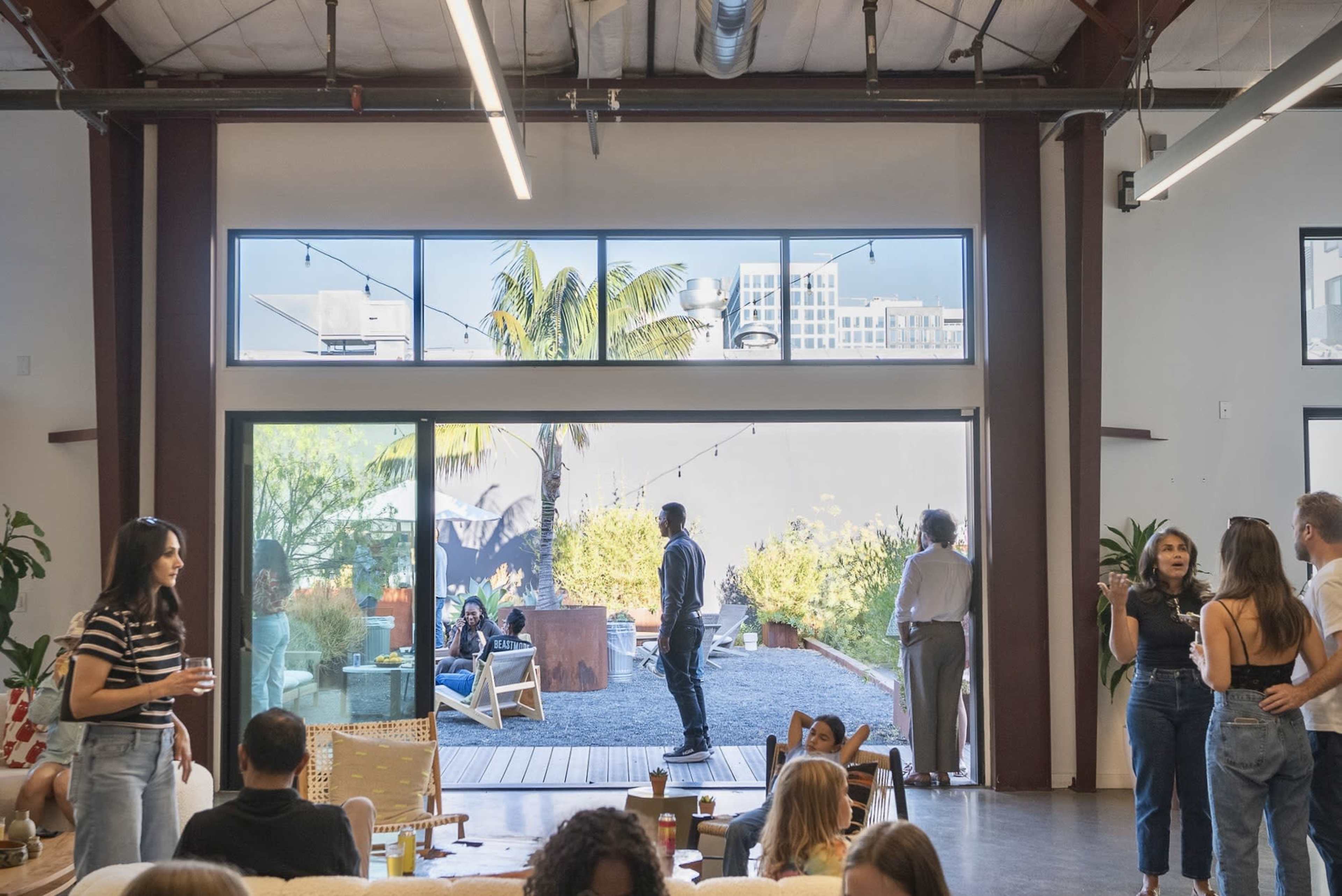 The image shows a modern indoor gathering space with people socializing, large windows overlooking an outdoor area with greenery, and a relaxed atmosphere.