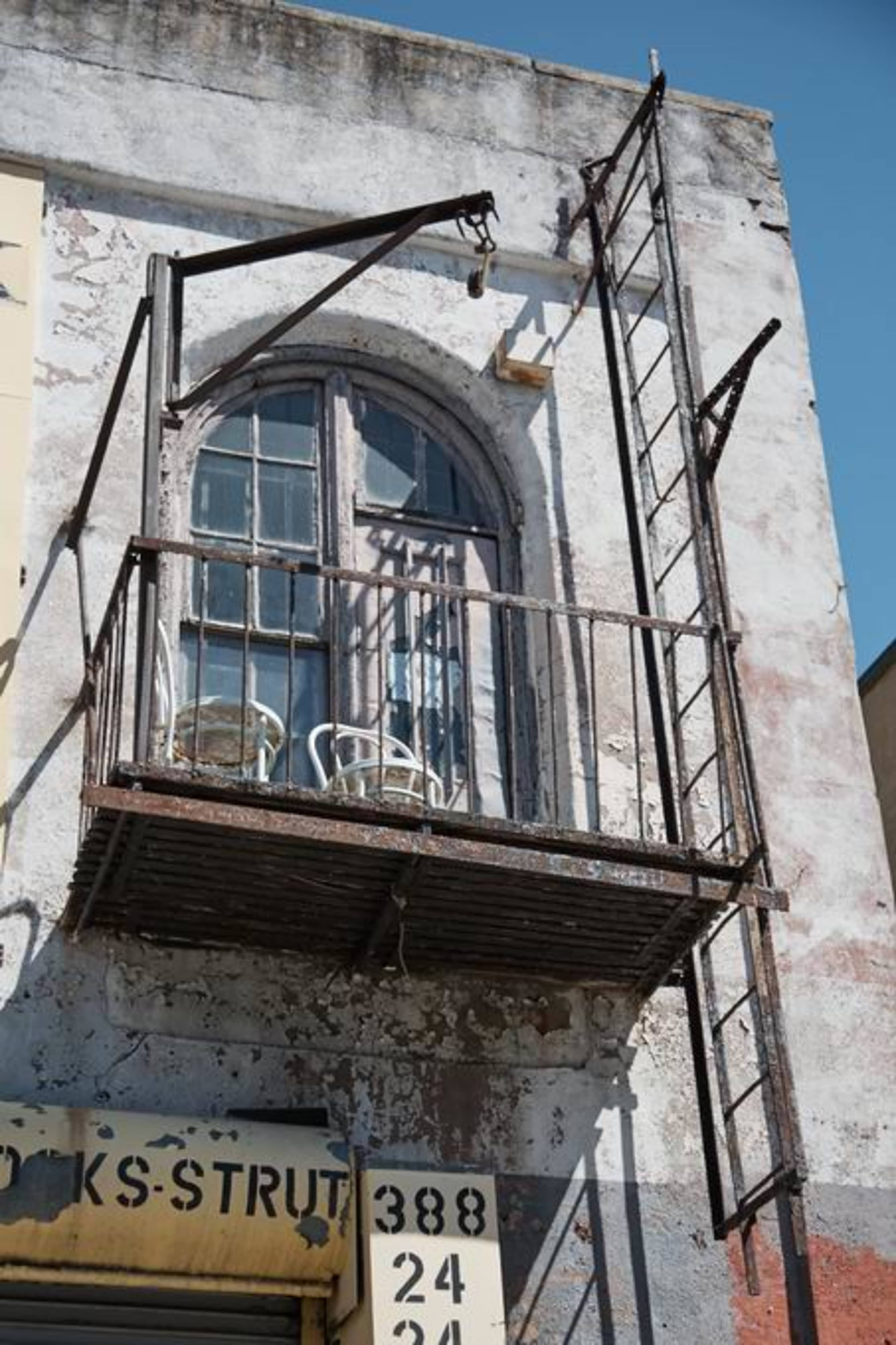 A weathered metal balcony with a ladder extends from an old brick building, featuring two white chairs on its surface.