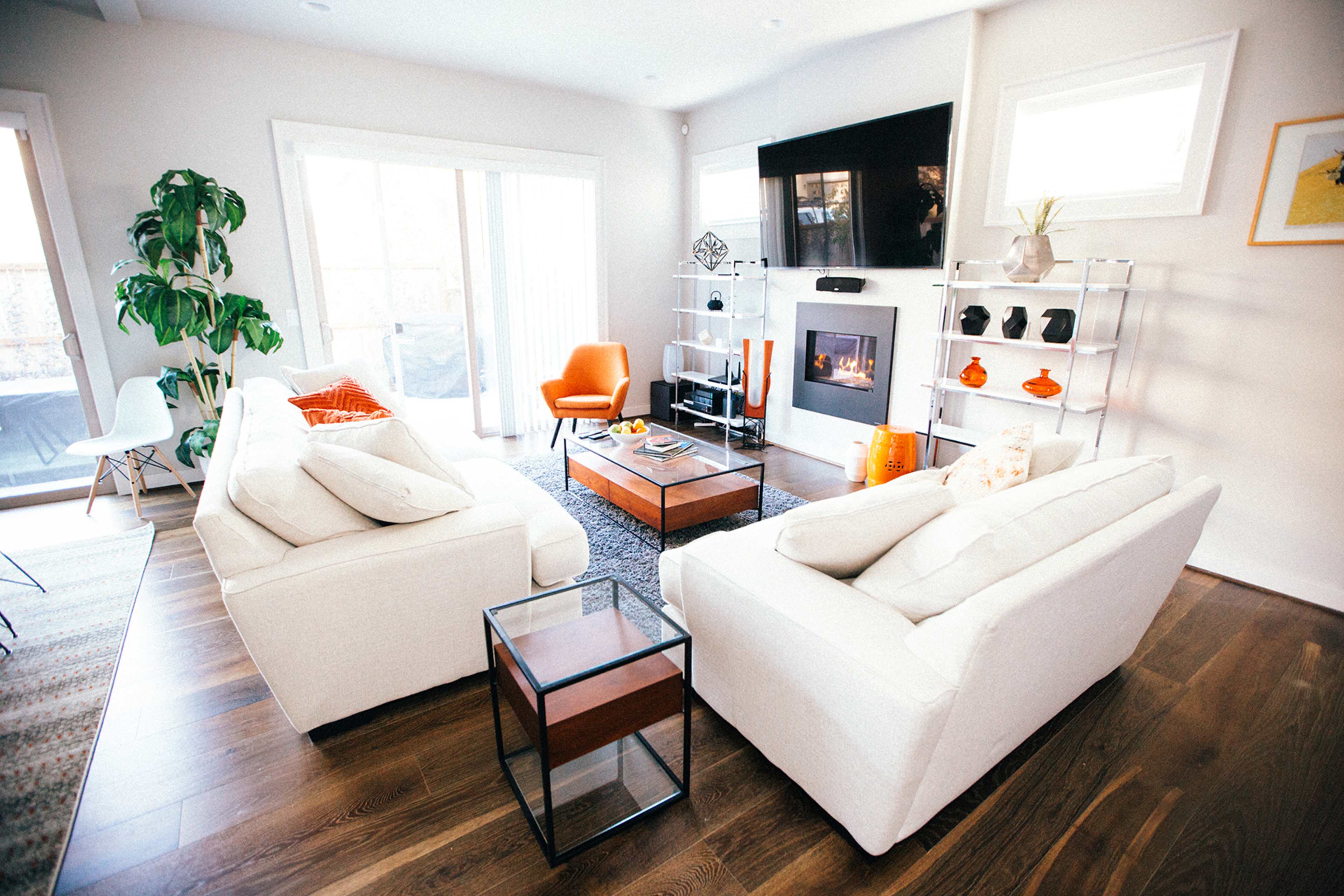 A modern living room features two white sofas facing a coffee table, a sleek fireplace with a television above, and large windows allowing natural light.