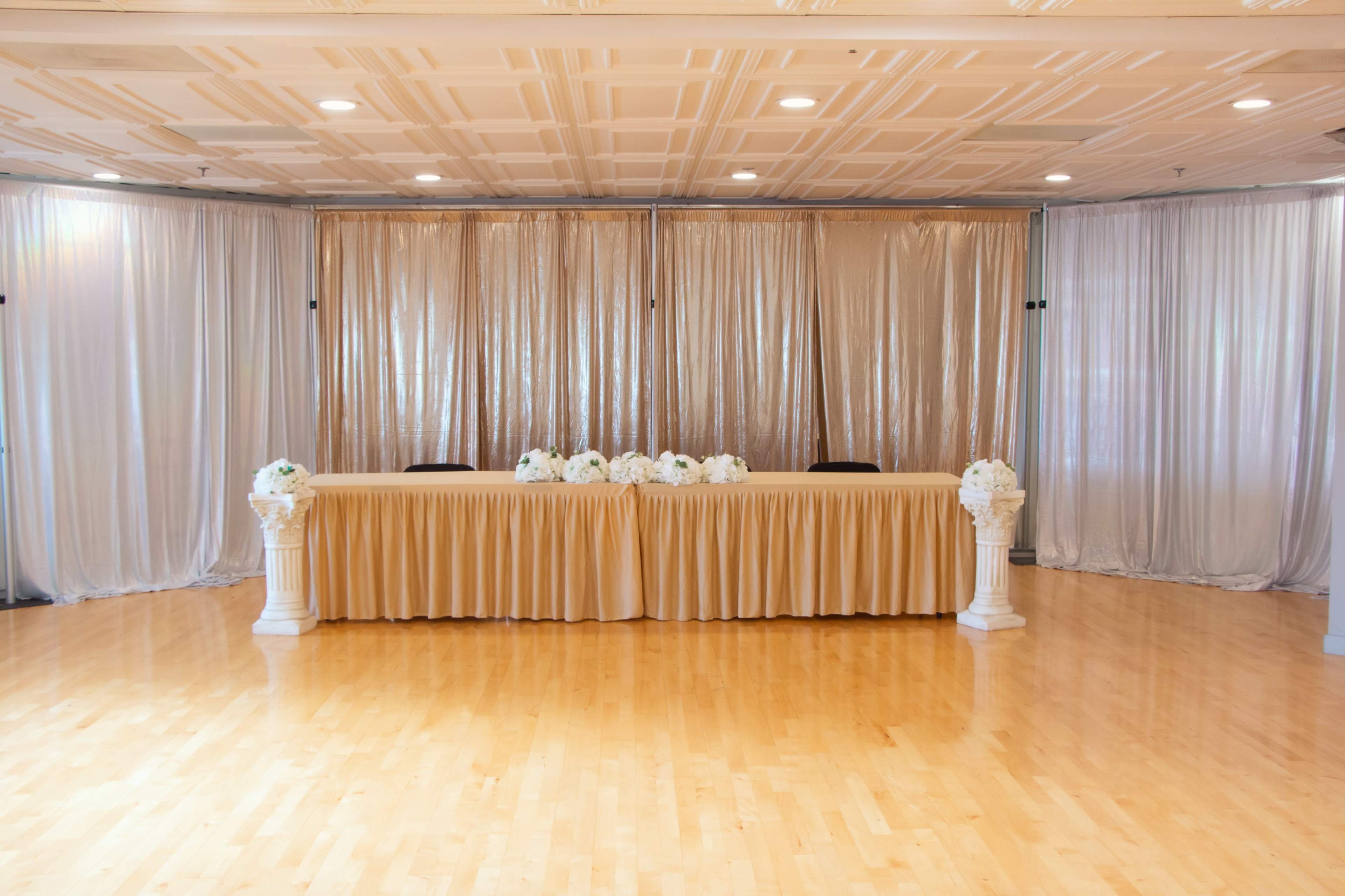 A decorated event space features a long table with a gold tablecloth, white floral arrangements, and sheer curtains in the background.