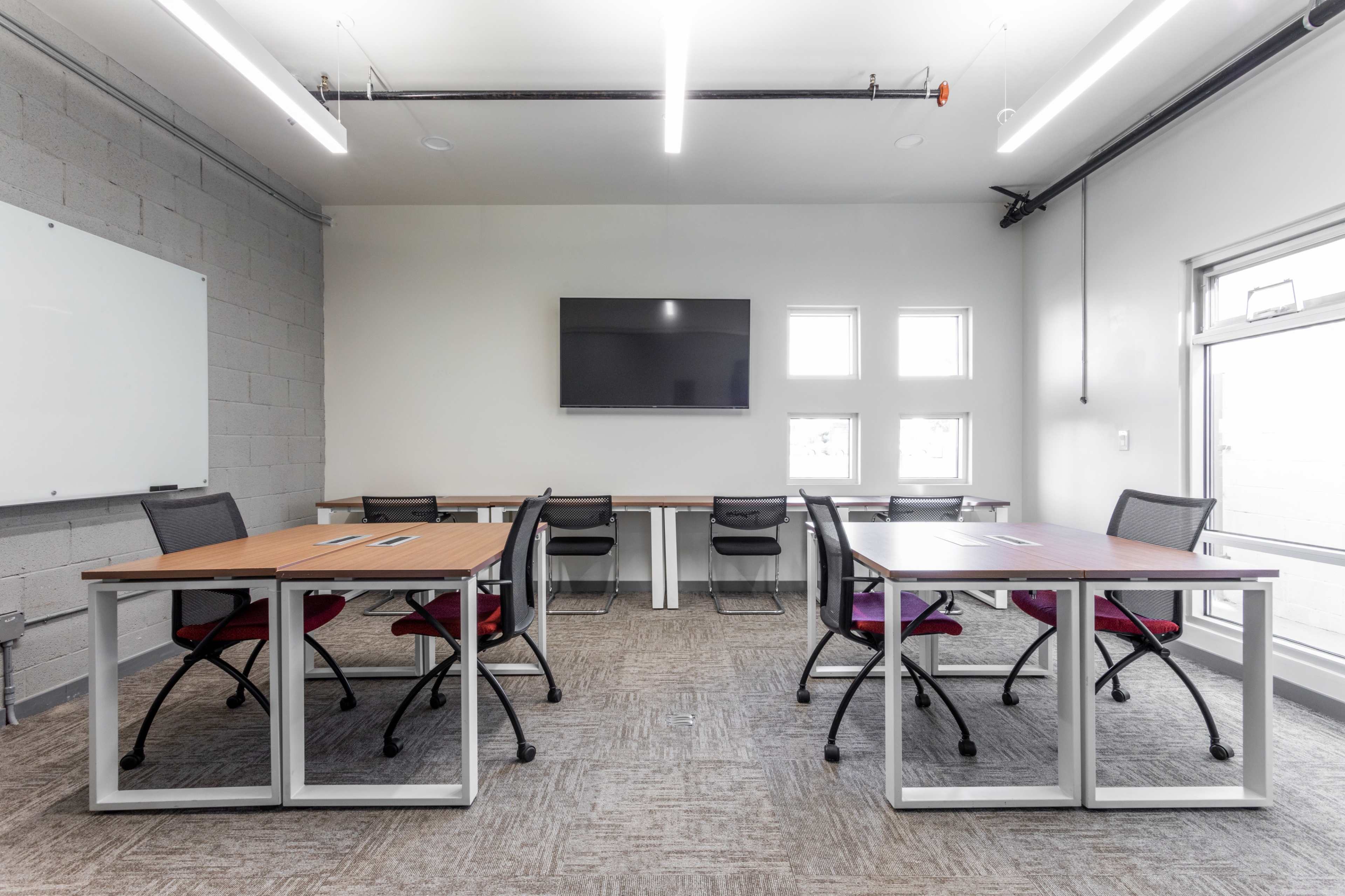 A modern office space featuring two sets of desks with chairs, a television mounted on the wall, and several windows.