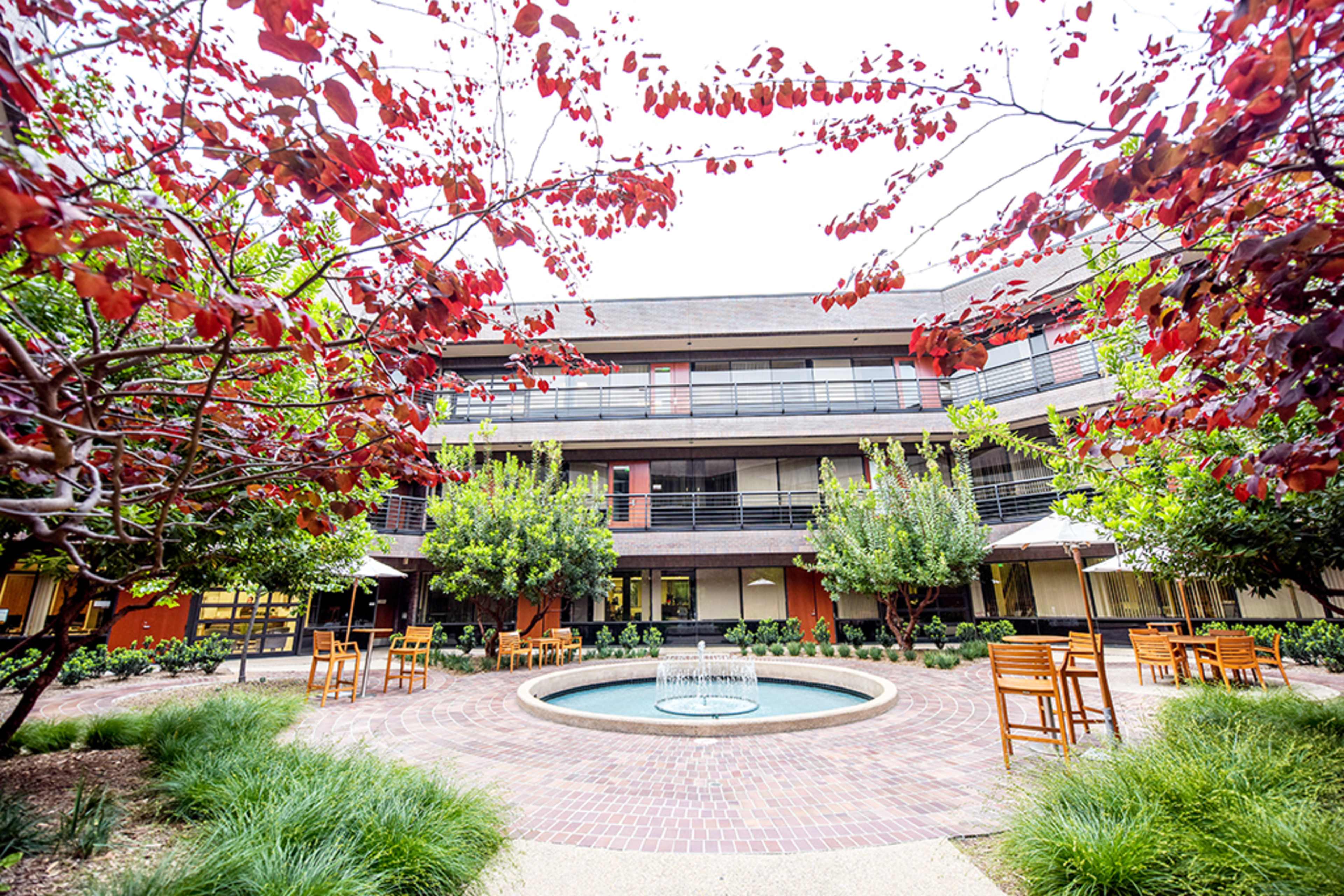 A courtyard features a central fountain surrounded by trees and seating in front of a three-story building.