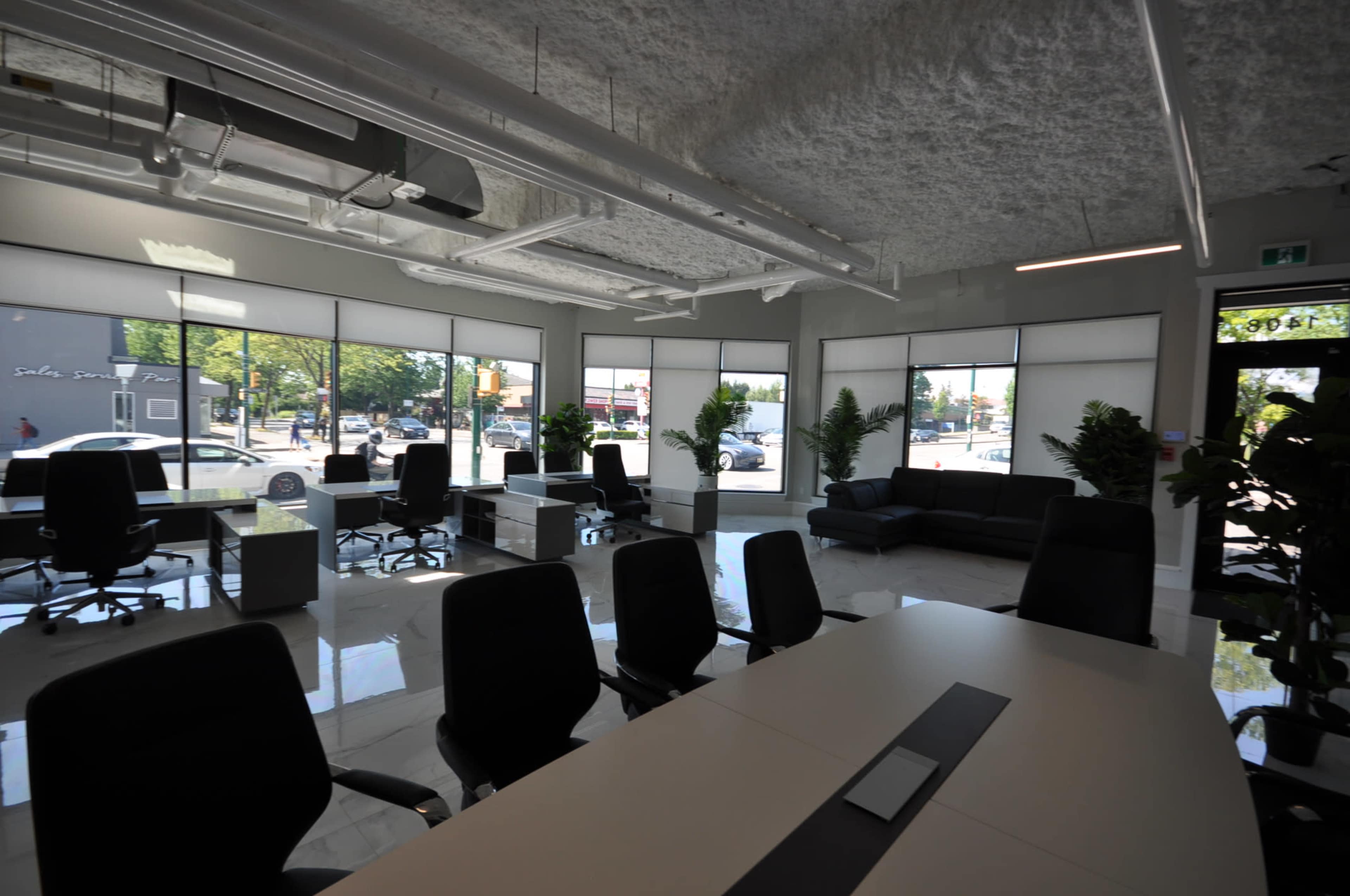 The image shows a modern office space featuring several desks, black chairs, large windows, and indoor plants.