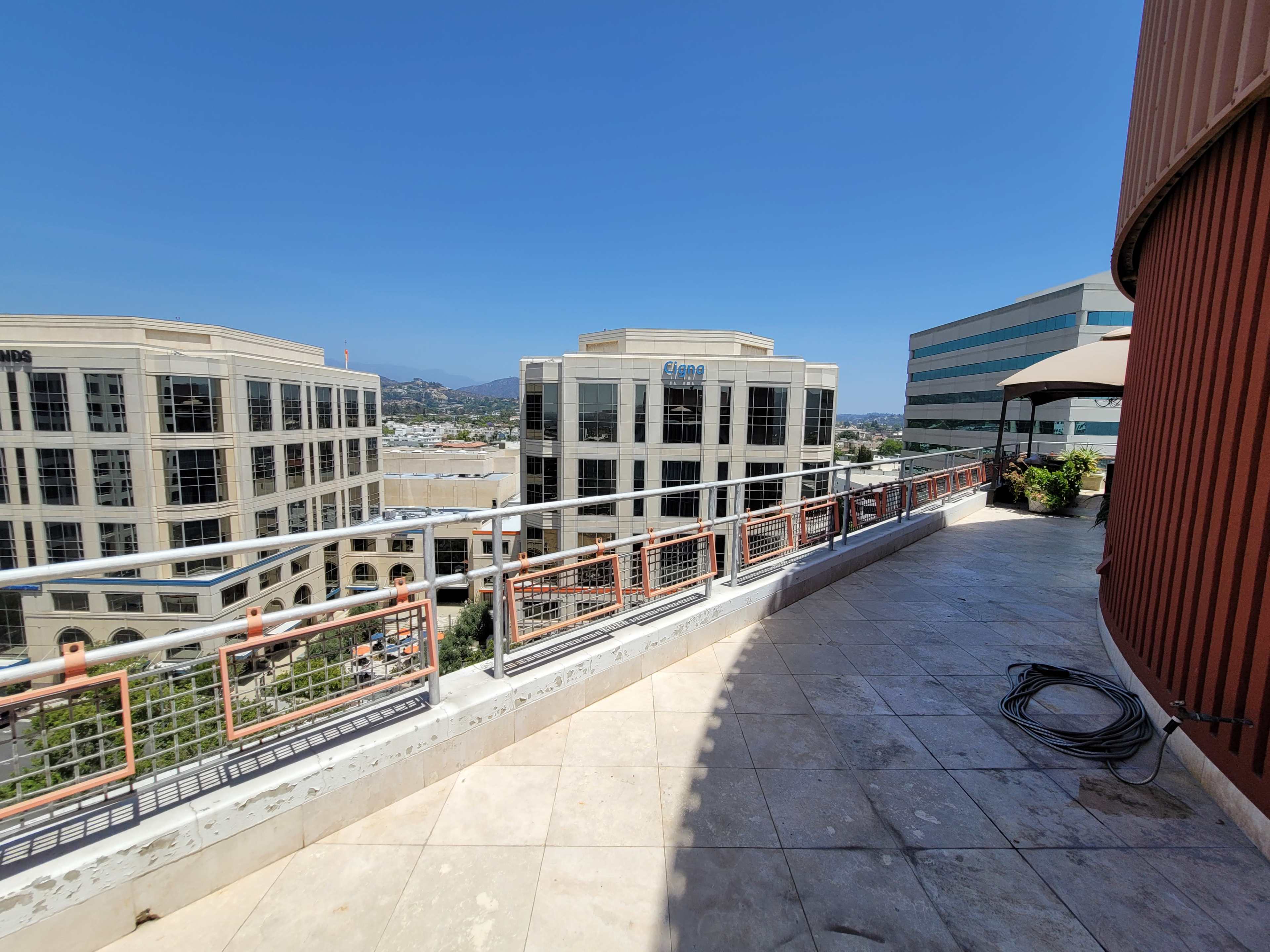 A sunlit balcony with tile flooring and a railing overlooking a row of commercial buildings and a clear blue sky.