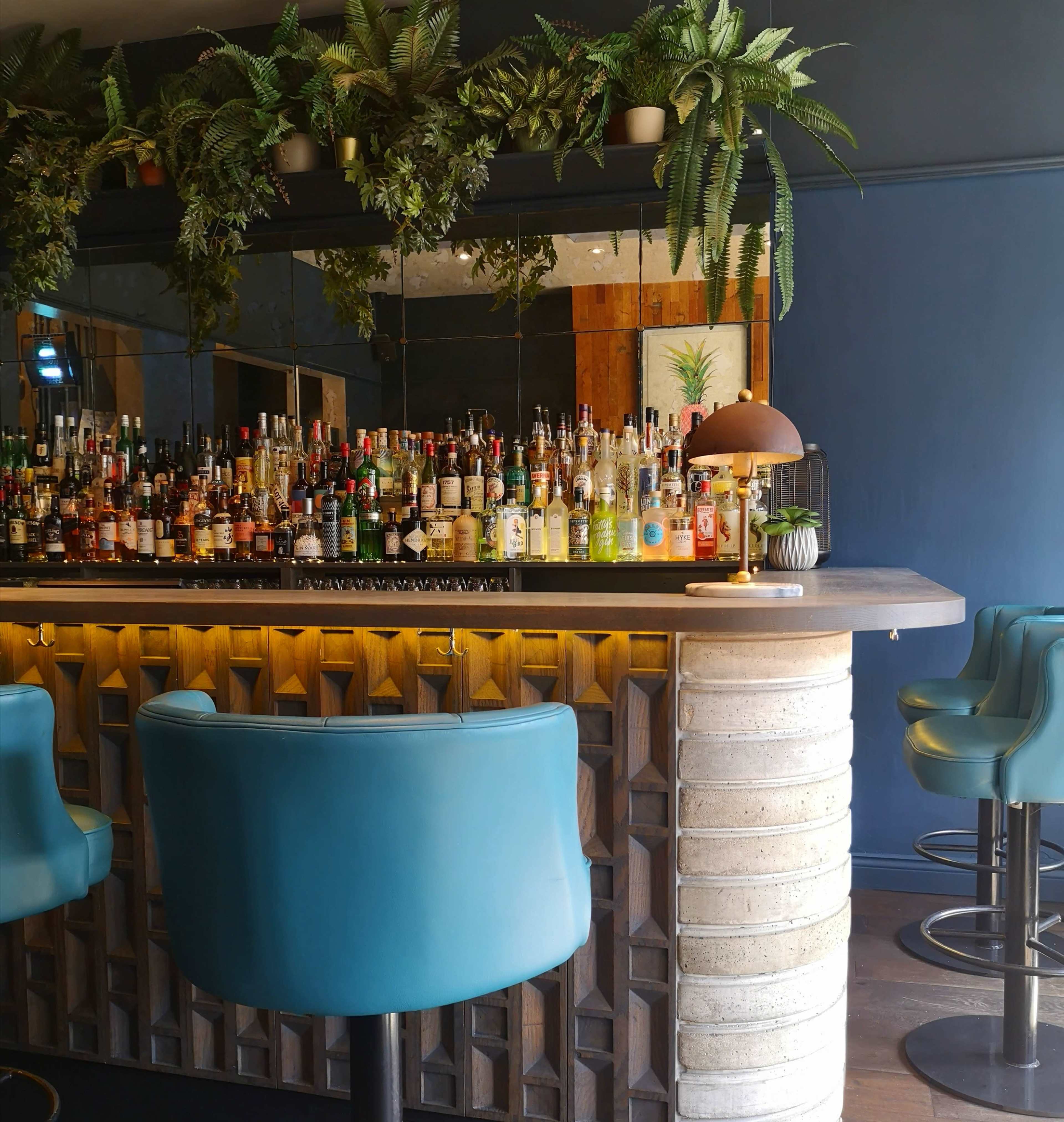 The bar features a variety of liquor bottles displayed on shelves behind a curved counter, with green plants adorning the walls and mirrored sections.