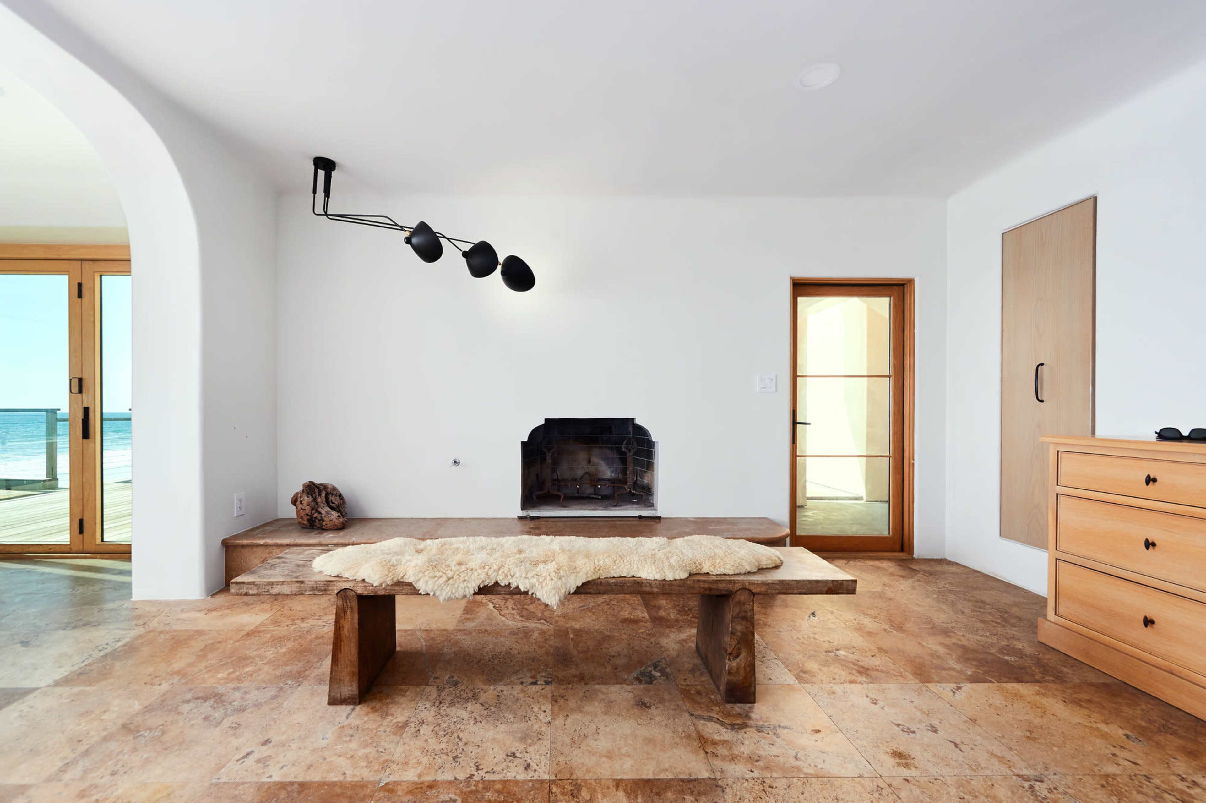 A minimalist living space featuring a wooden table with a fur throw, a fireplace, and a large window that offers a view of the ocean.