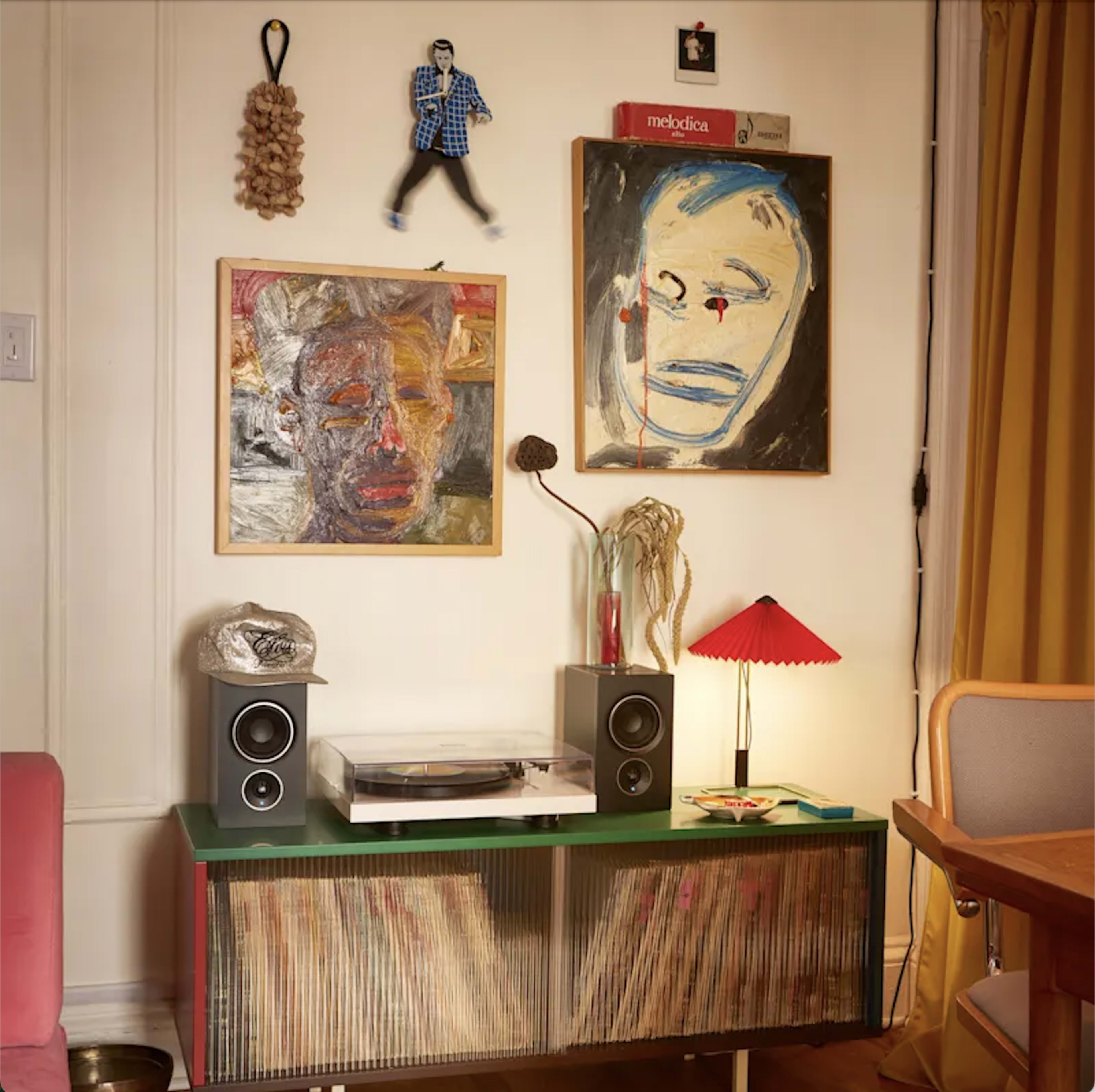 The image shows a corner of a room featuring a wooden record player stand with vinyl records, two speakers, and colorful artwork on the wall above.