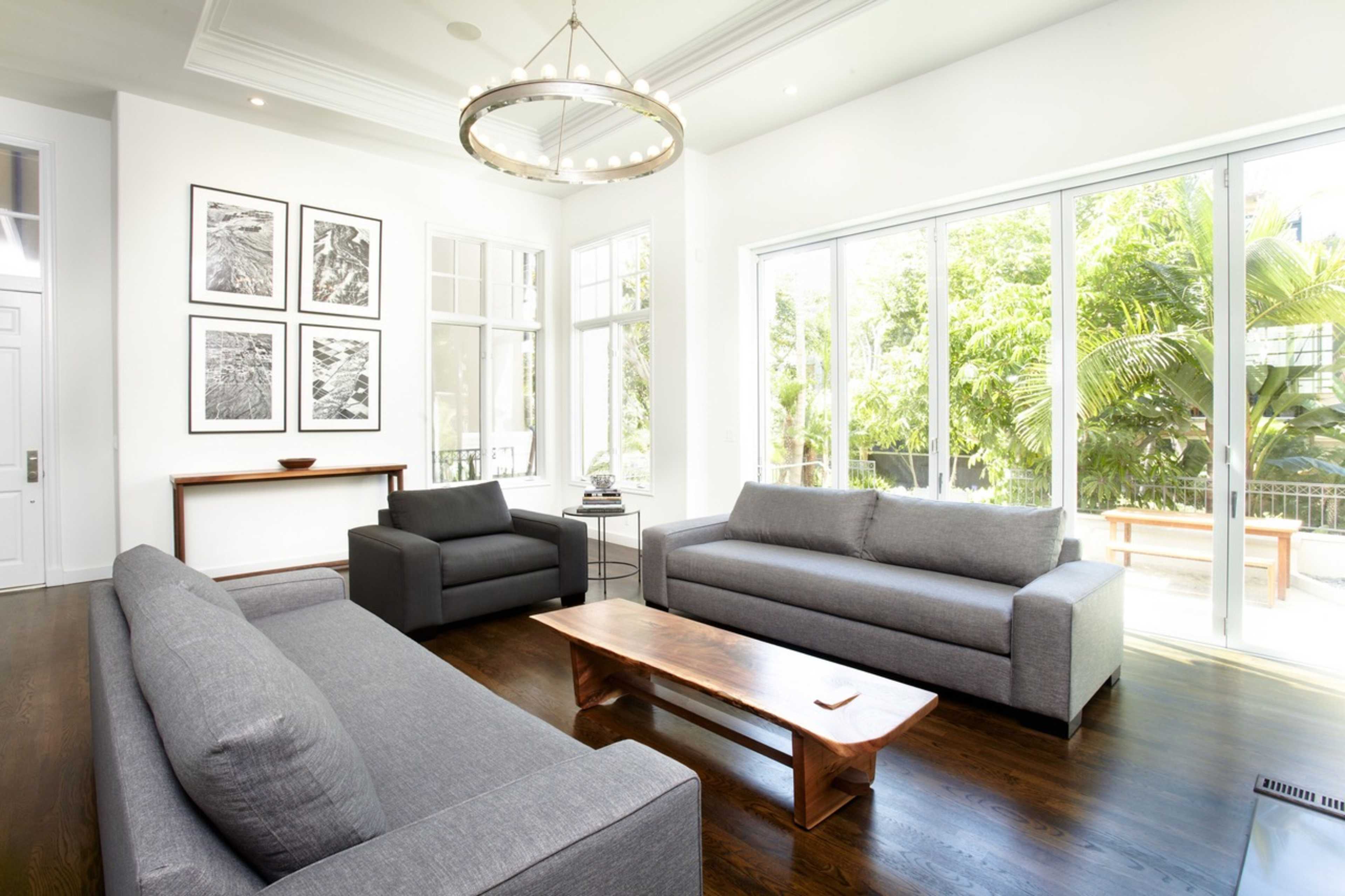 A modern living room features gray sofas, a wooden coffee table, and large windows overlooking lush greenery.