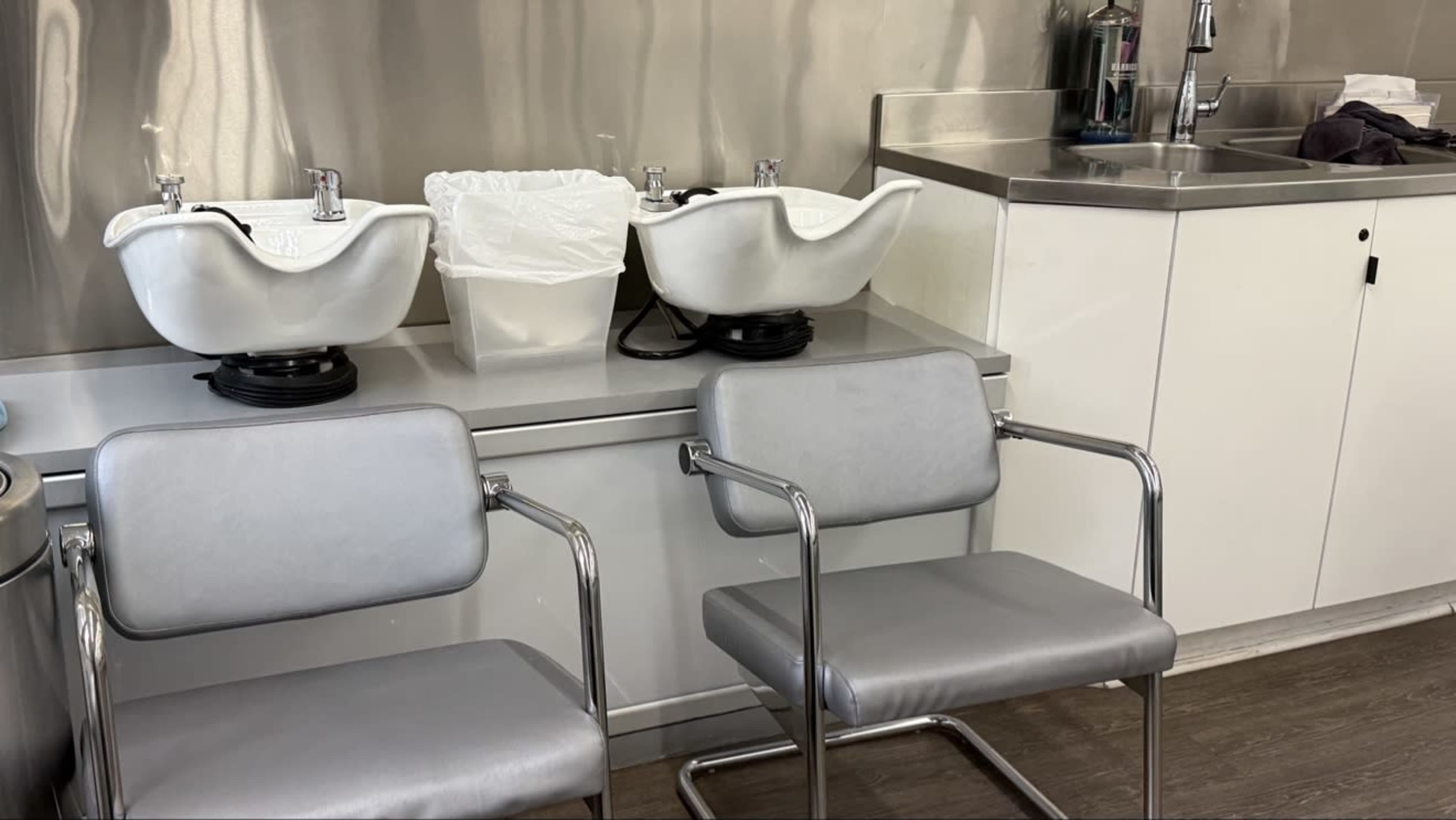 The image shows two salon shampoo bowls next to a sink, with two silver chairs positioned in front of them.