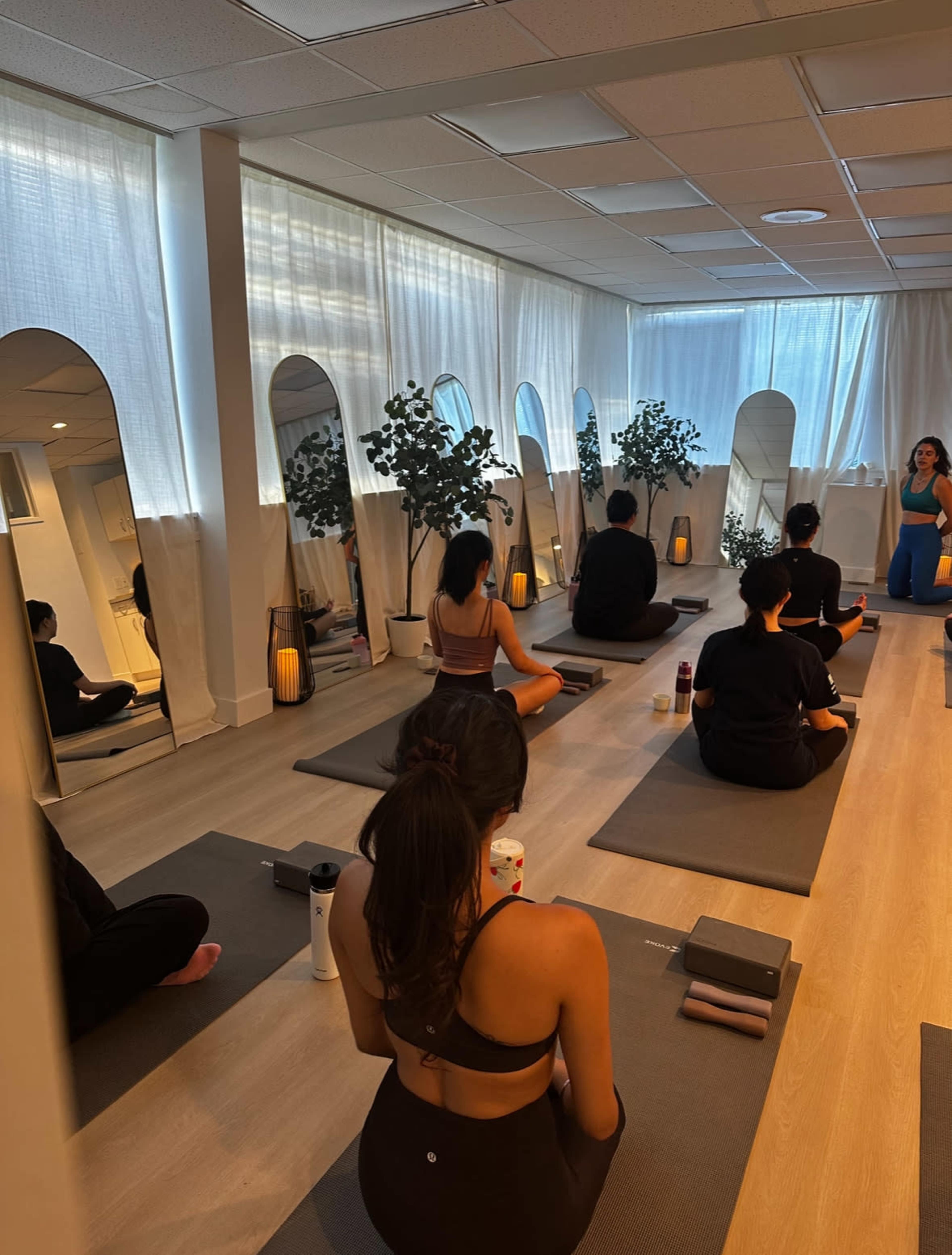 A group of individuals is seated on yoga mats in a well-lit studio with mirrors and plants, preparing for a class.