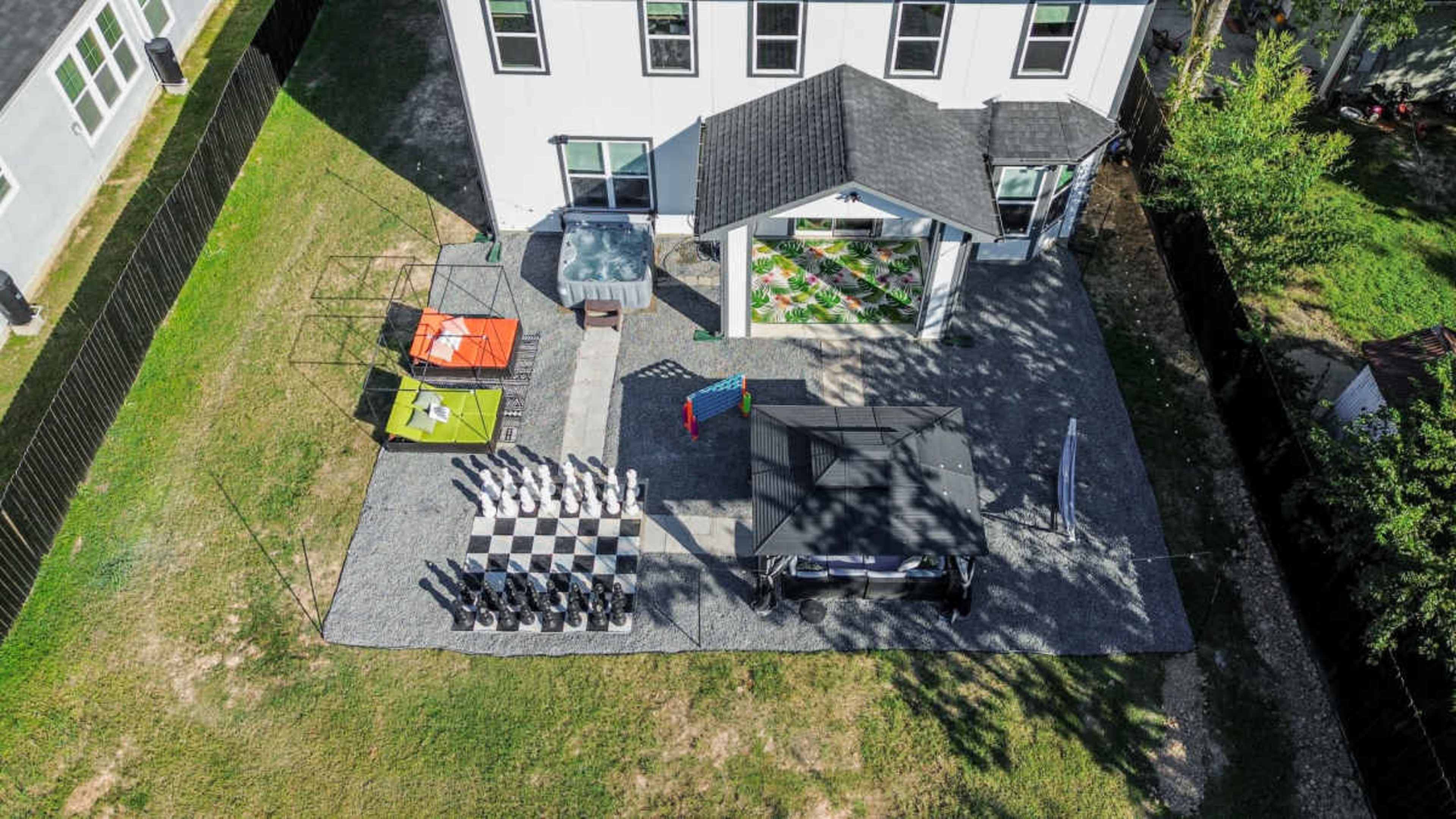 The image shows a backyard with a large outdoor chess set, a hot tub, colorful lounge furniture, and a covered seating area next to a house.