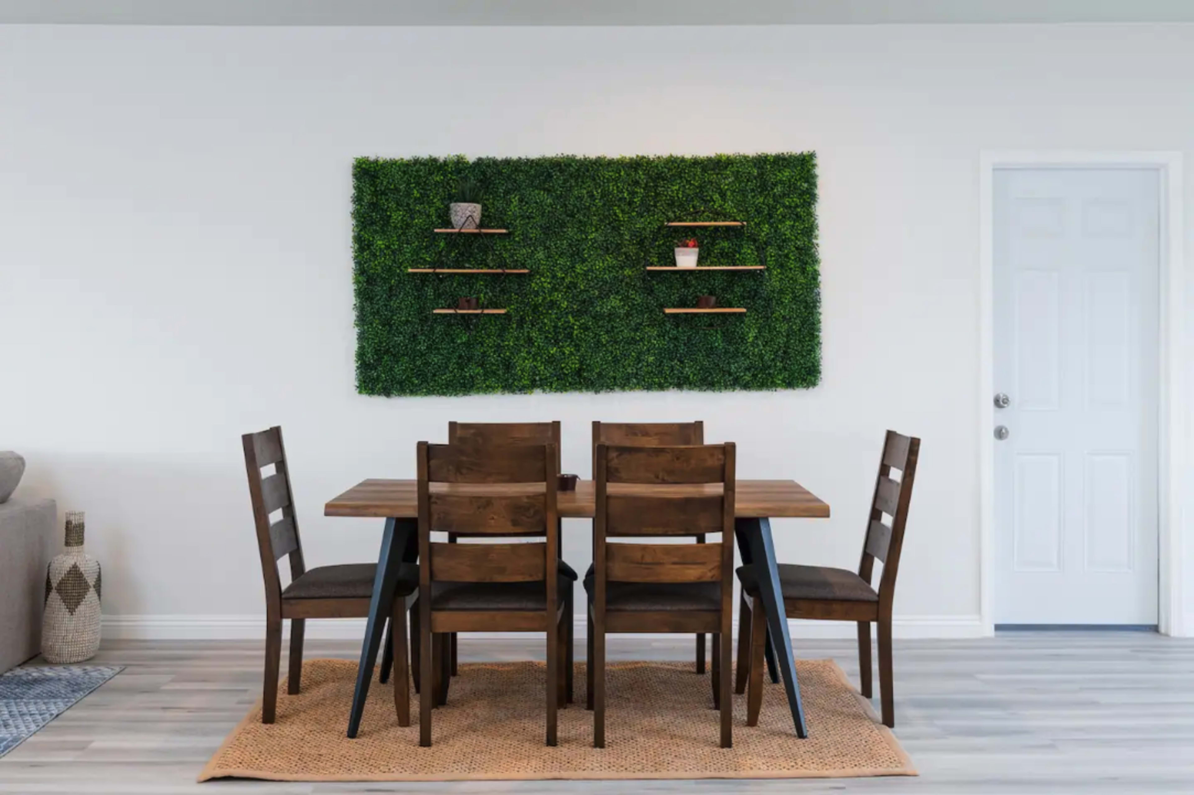 A wooden dining table with four chairs is set against a wall featuring a green artificial plant display and shelves, with a door visible to the right.