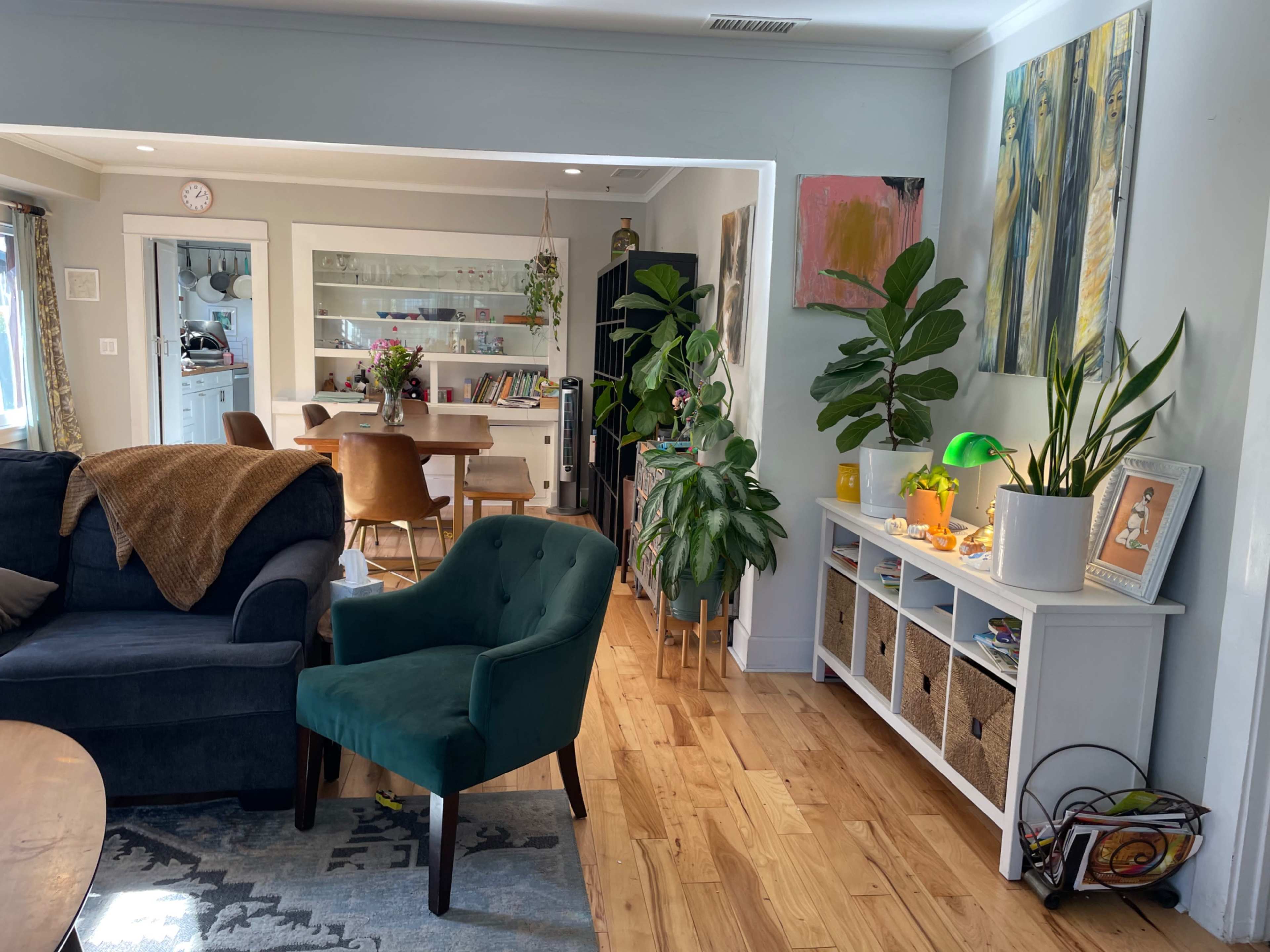 The image shows a living room connected to a dining area, featuring a blue sofa, a green chair, potted plants, and a white storage unit filled with baskets.