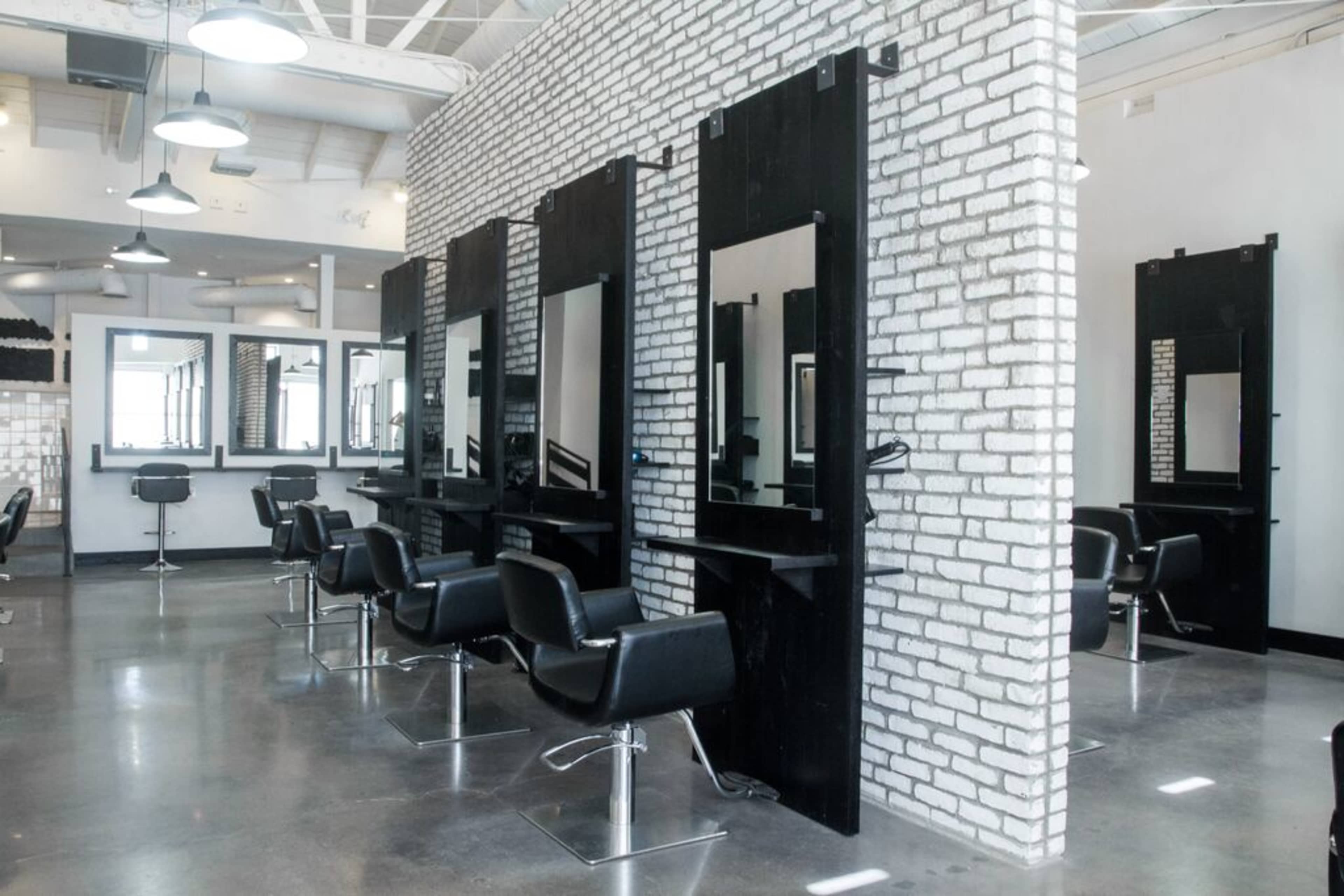 The image shows a modern hair salon with black chairs and mirrors against a white brick wall.