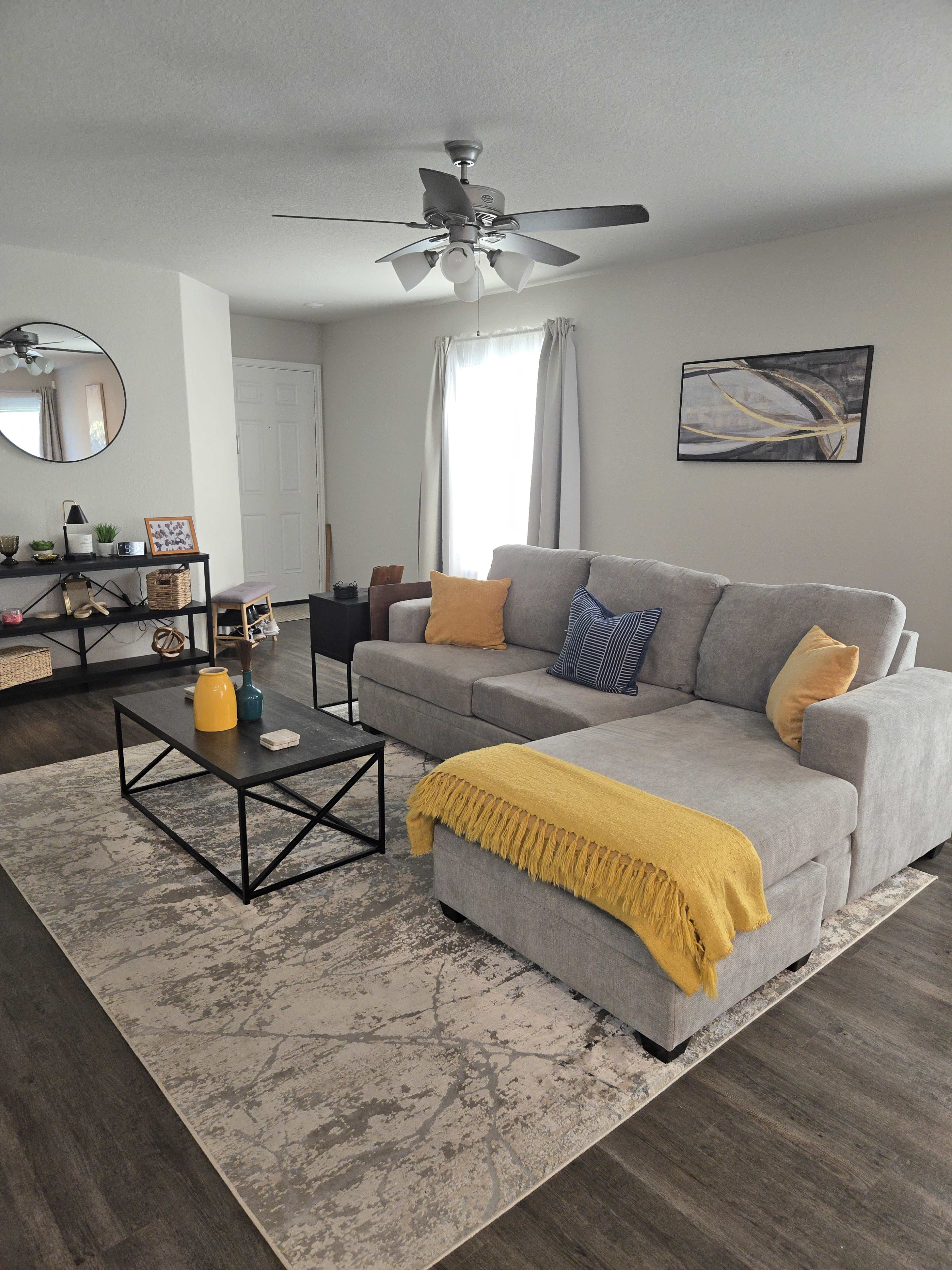 The image shows a modern living room featuring a gray sectional sofa with a yellow throw blanket, a coffee table, and a round mirror on the wall.